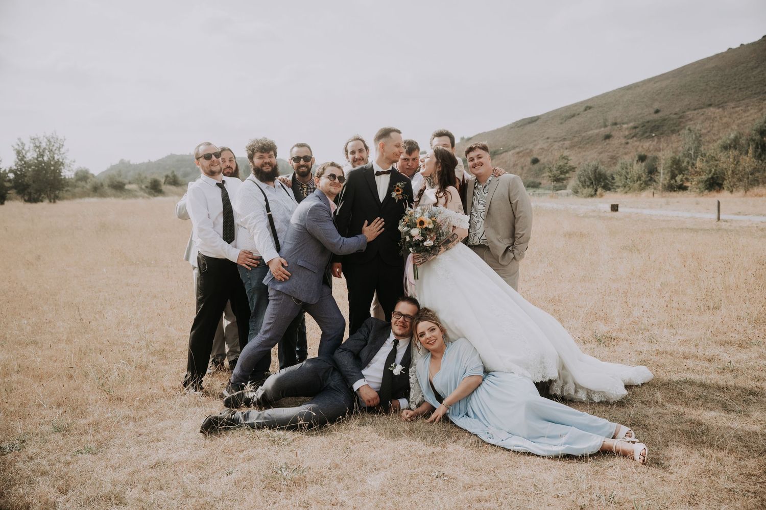 Wedding party posing playfully in a golden field, bride and groom surrounded by groomsmen and bridesmaids.