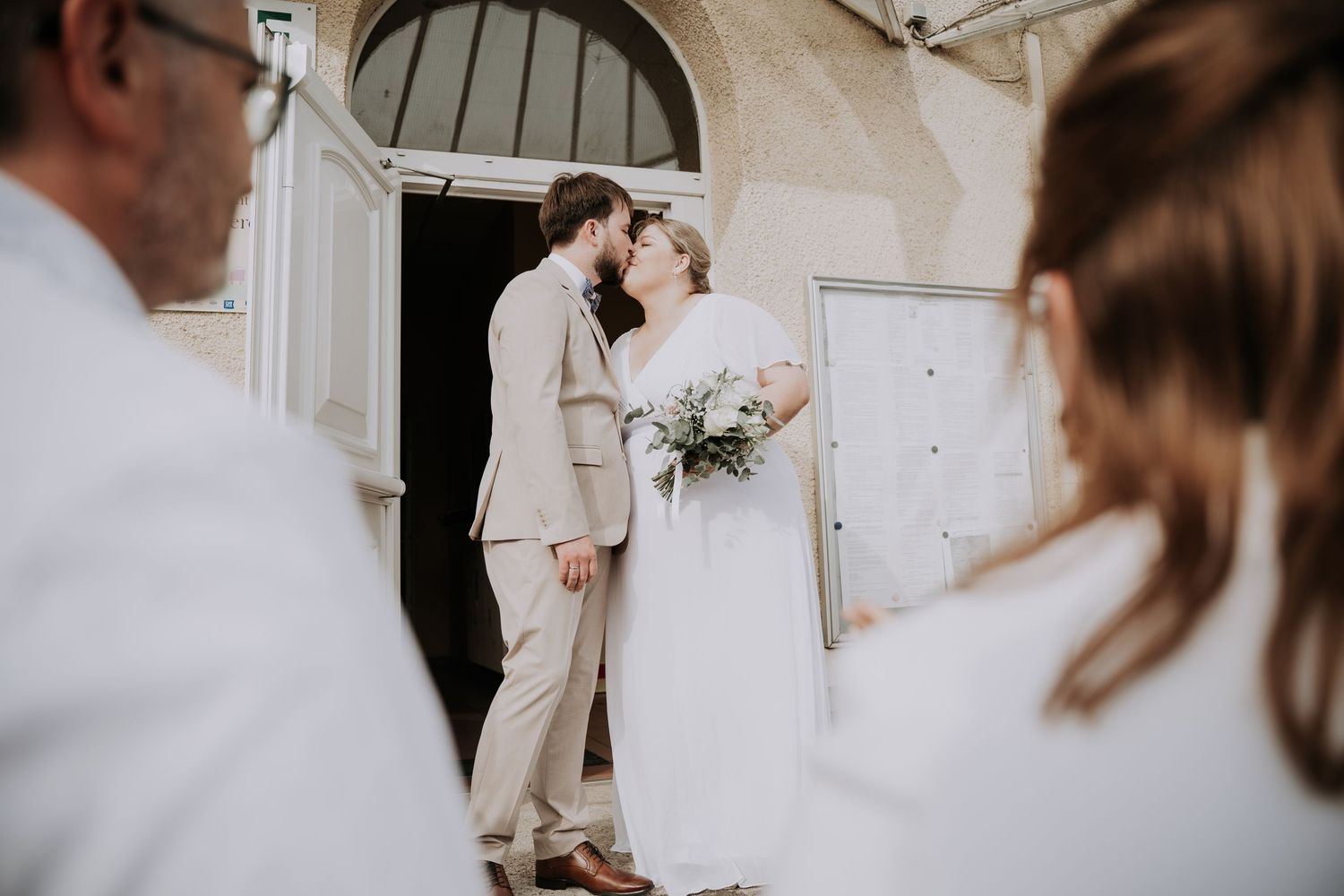 Bride and groom share a kiss outside a church, bride holding a white bouquet, guests in foreground.