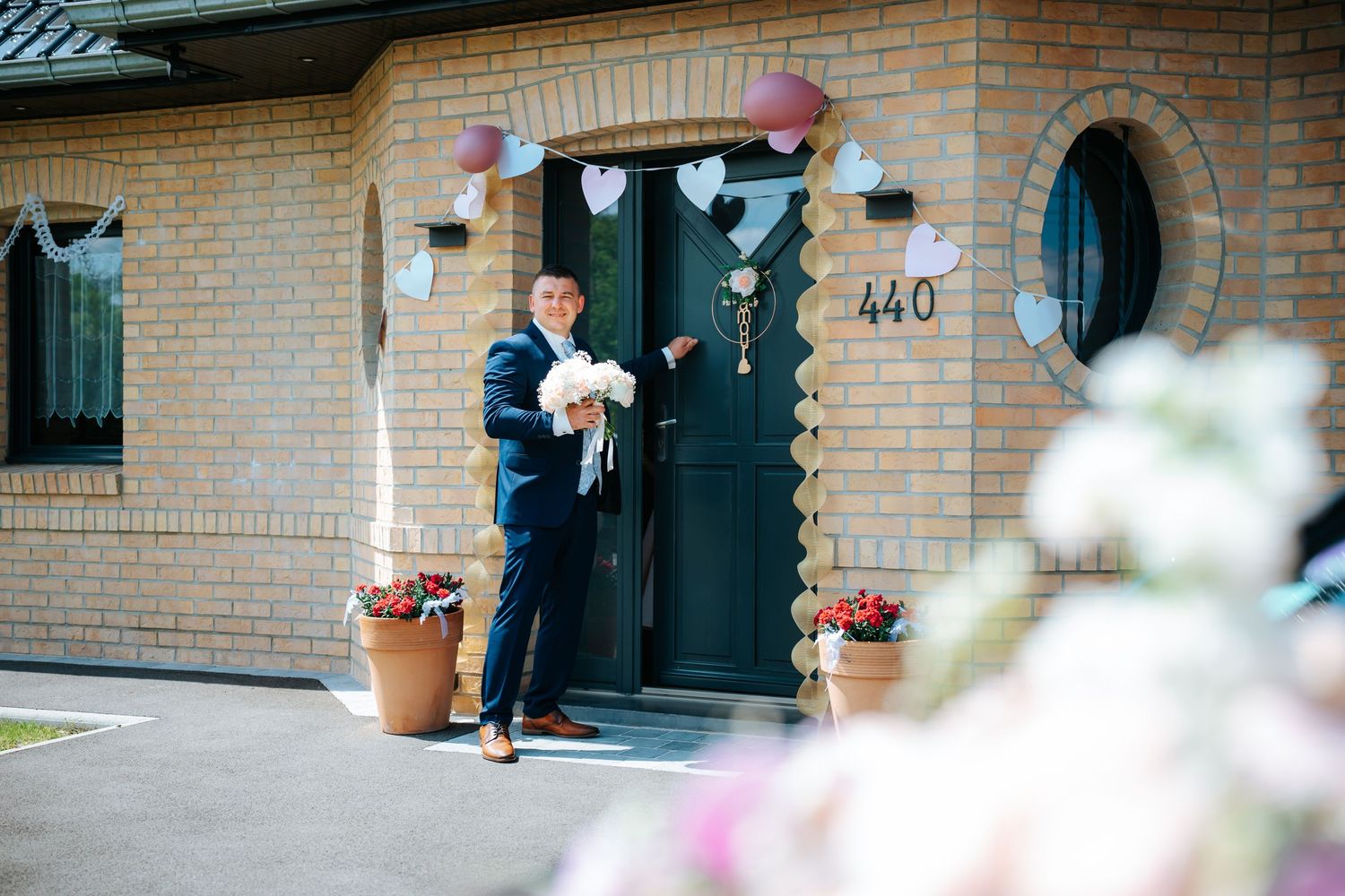 Groom in navy suit holding flowers stands at decorated brick house entrance numbered 440 with heart bunting for wedding.