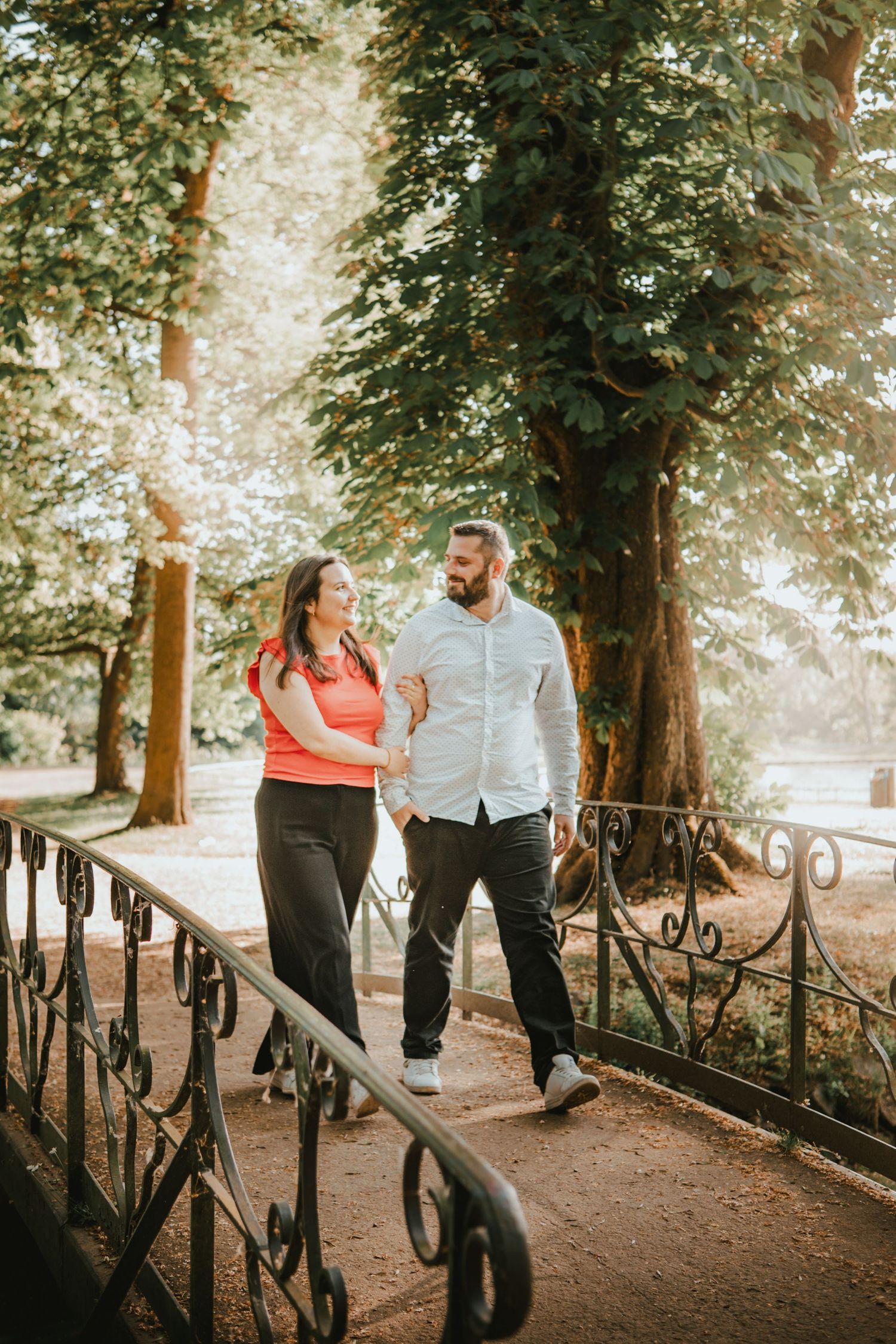 Couple walking hand in hand through a sunlit park path, smiling at each other near ornate iron railings.