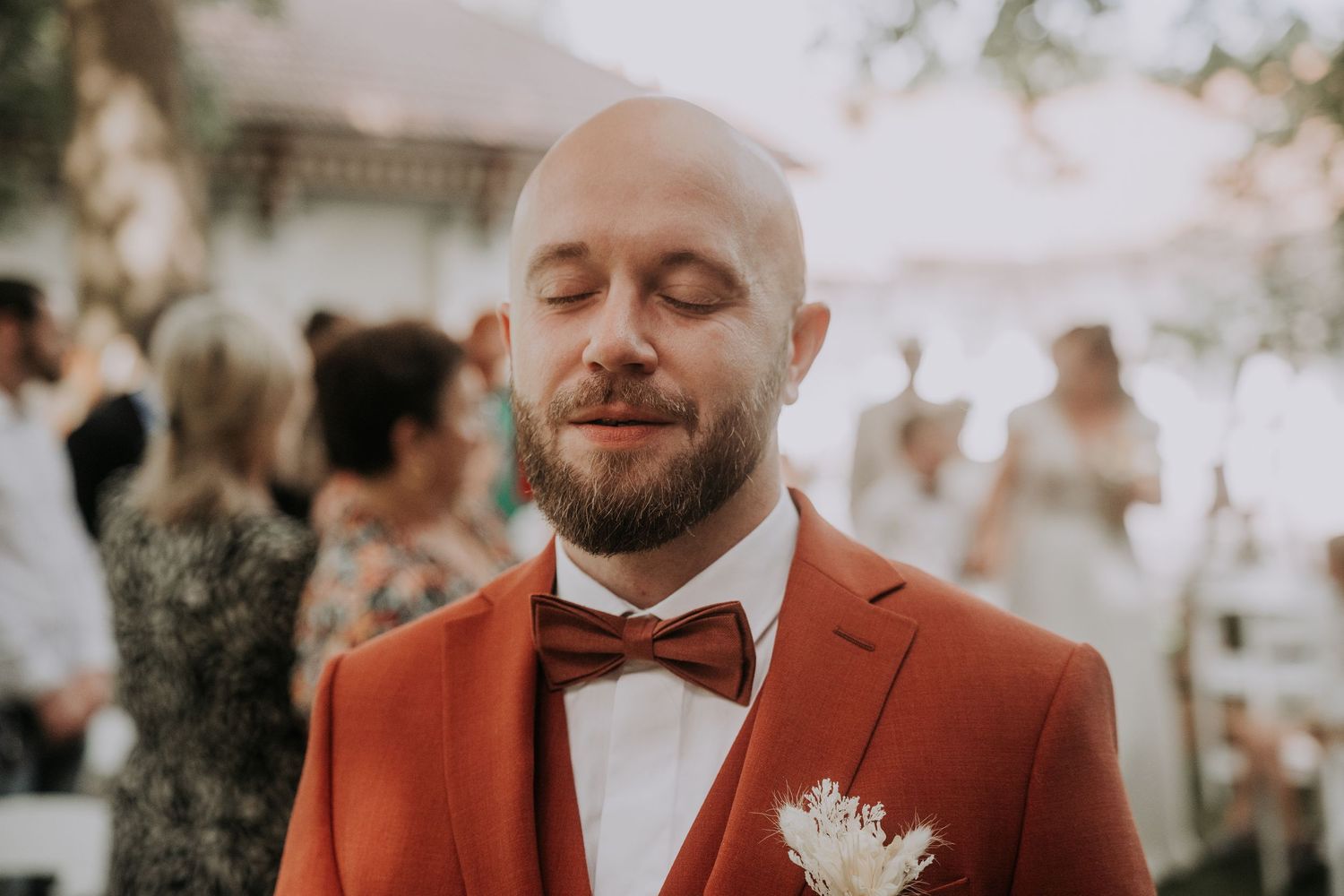 Bald groom with beard wearing rust orange suit and bow tie at outdoor wedding ceremony.