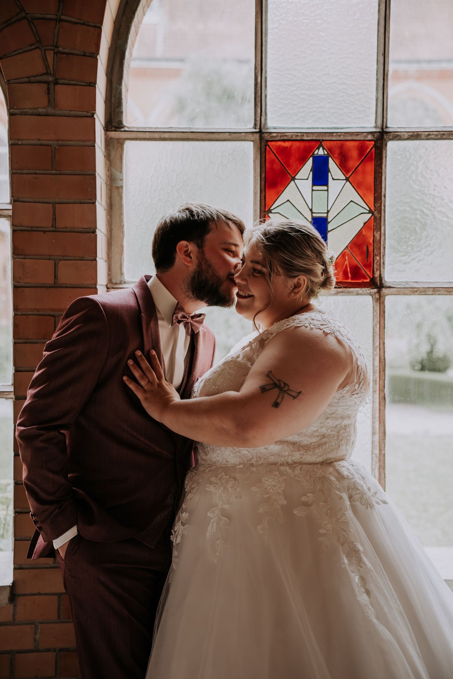 A bride and groom share a romantic moment by a stained glass window, with the groom kissing her cheek in a historic brick venue.