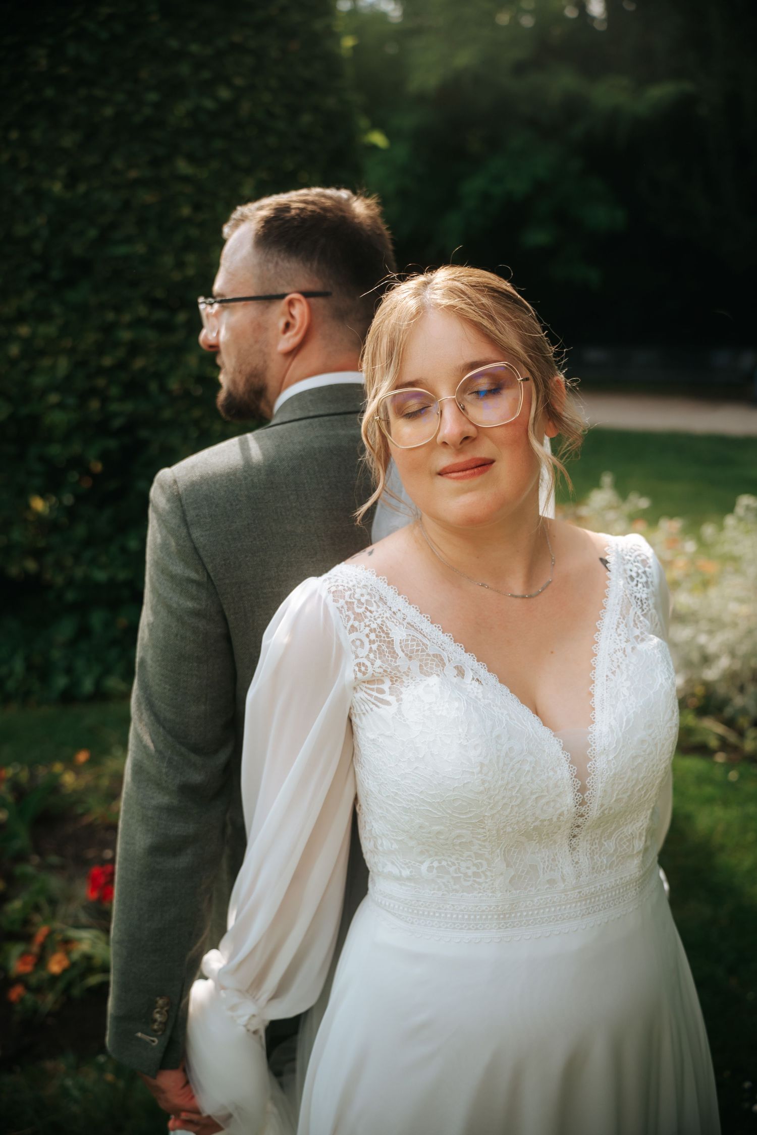 Bride in white lace gown and glasses holds groom's hand in a lush garden during outdoor wedding portrait.