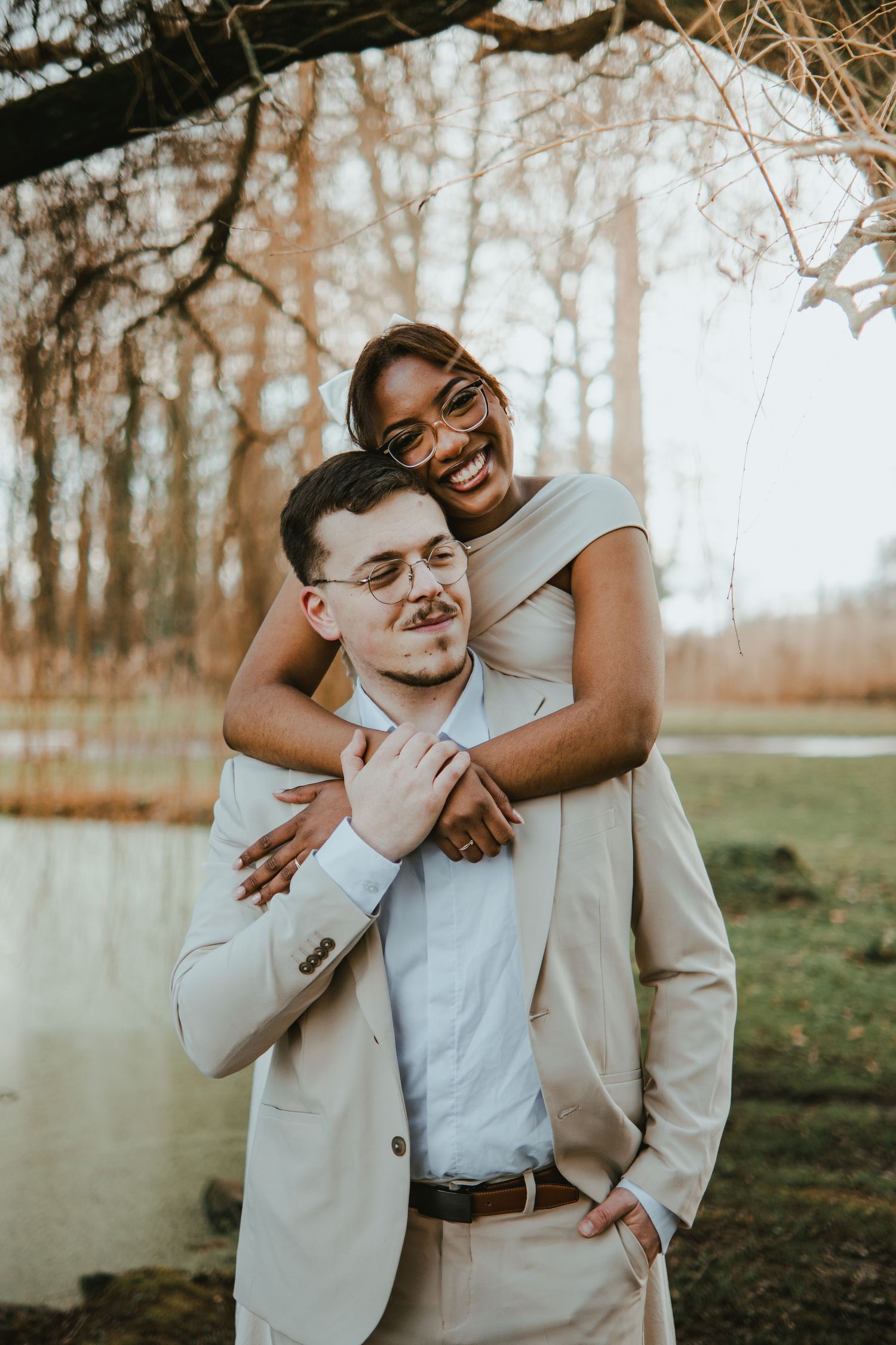 Smiling interracial couple in beige outfits embracing outdoors near a pond with bare trees in background.