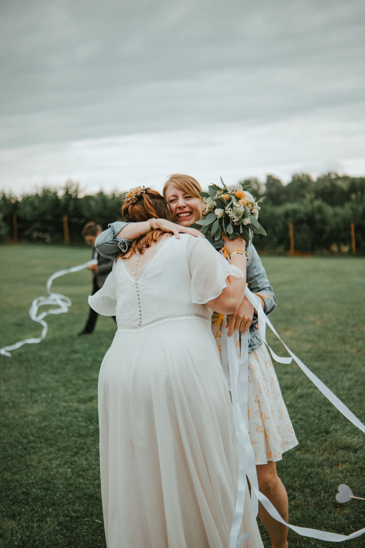 Bride in white dress hugging smiling guest holding bouquet outdoors at wedding reception on green lawn.