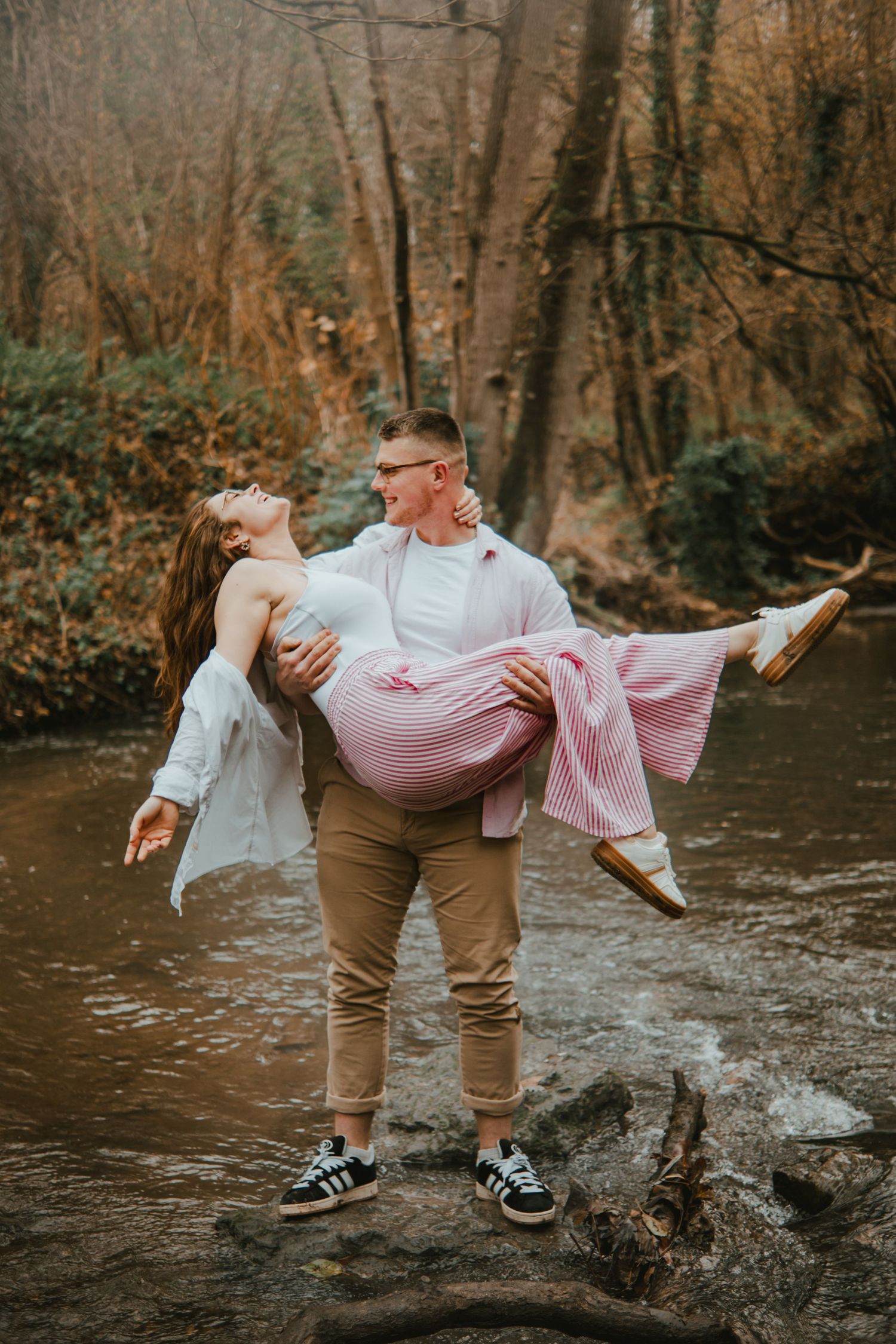 Man carrying woman in pink dress by a forest creek, romantic outdoor couple photo session.