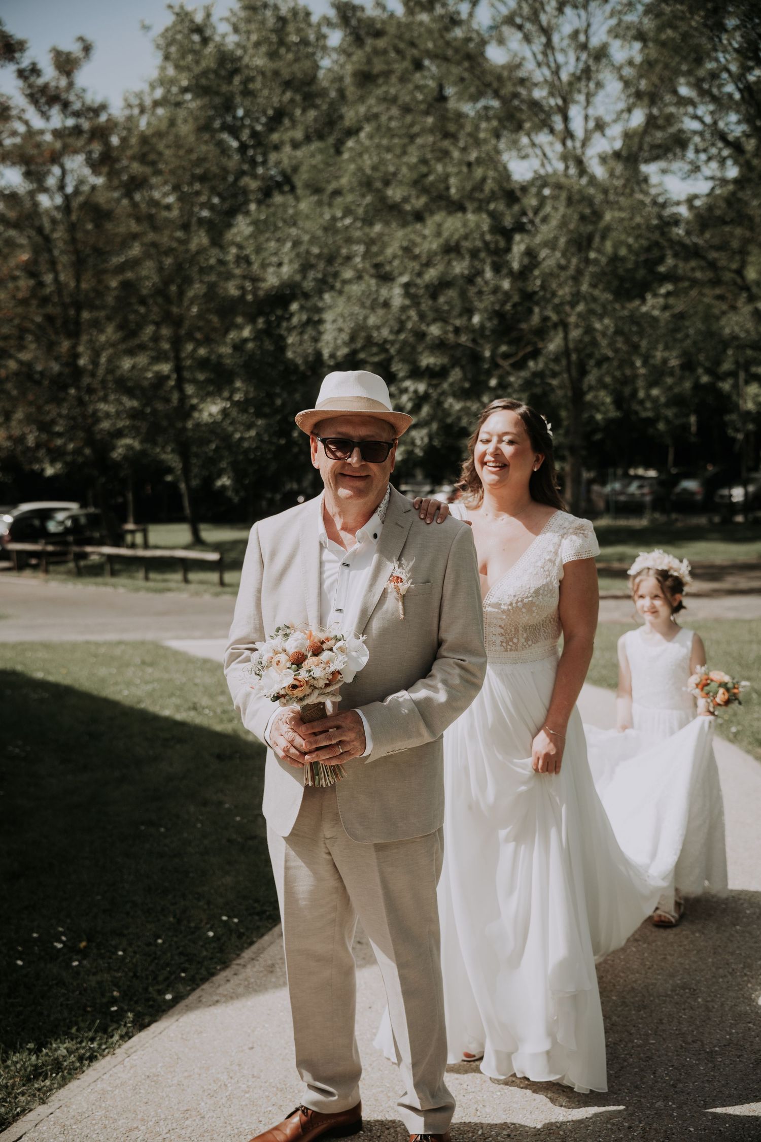 Bride walks with man in tan suit and hat holding bouquet, flower girl follows on sunny park path.