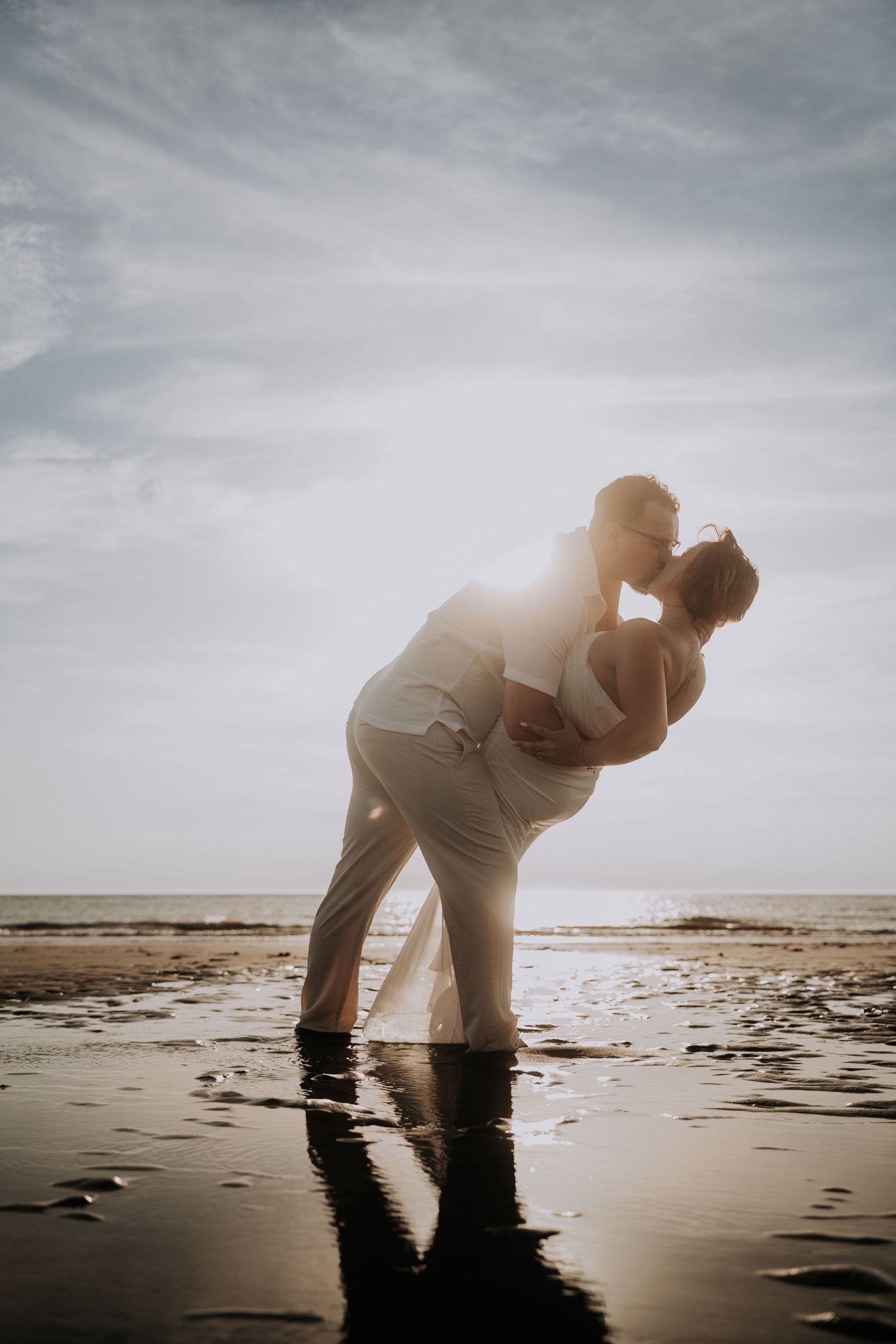 Couple sharing a romantic kiss on a reflective beach at sunset, silhouetted against a glowing sky.