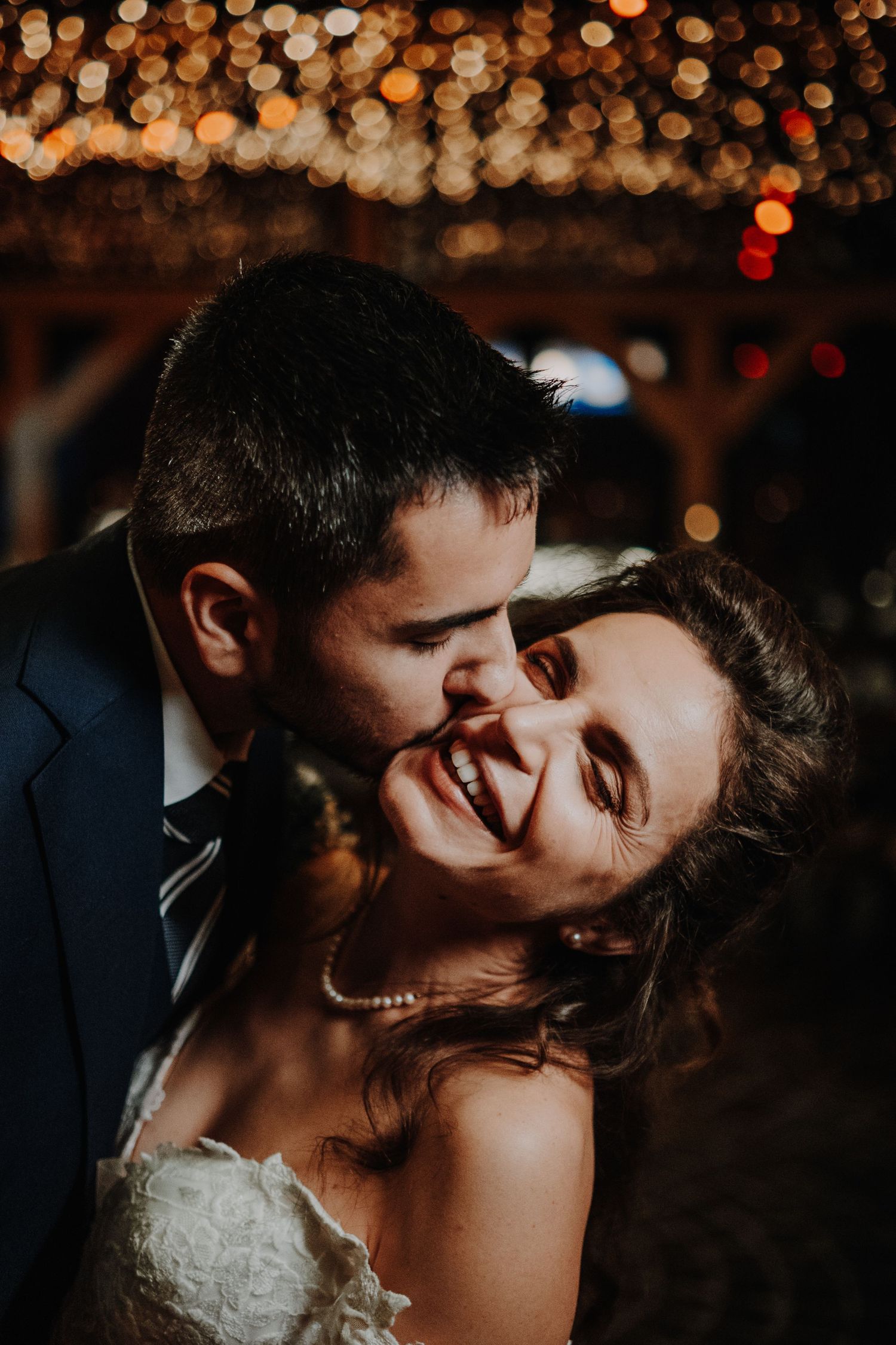 A groom kissing his smiling bride on the cheek under warm bokeh string lights at a romantic wedding reception.