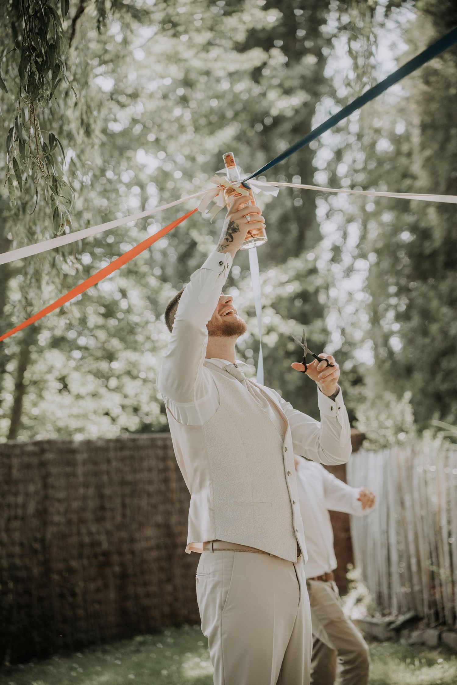 Man in white suit holding colorful ribbons outdoors at a garden celebration event.