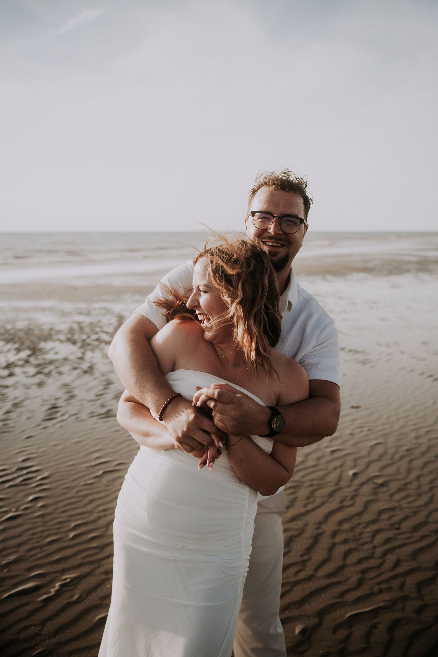 A couple embraces on a sandy beach, the woman in a white dress and the man in a white shirt, smiling joyfully.