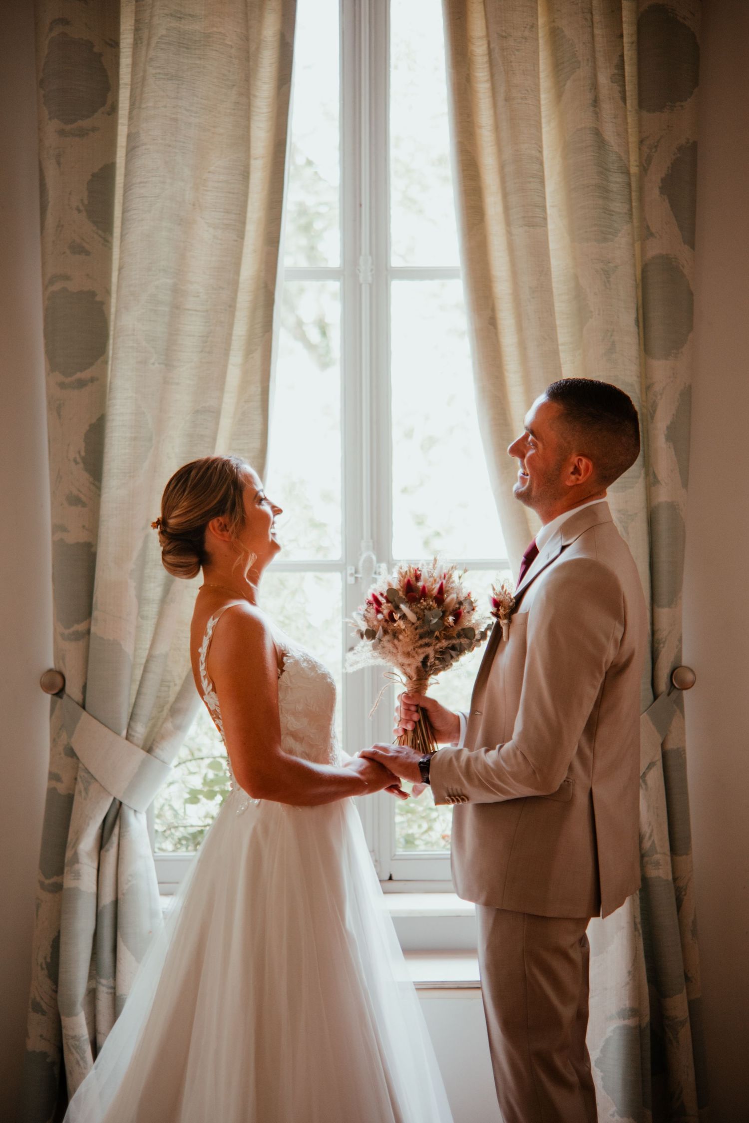 Bride and groom share a romantic moment by a sunlit window, holding a bouquet together in elegant wedding attire.