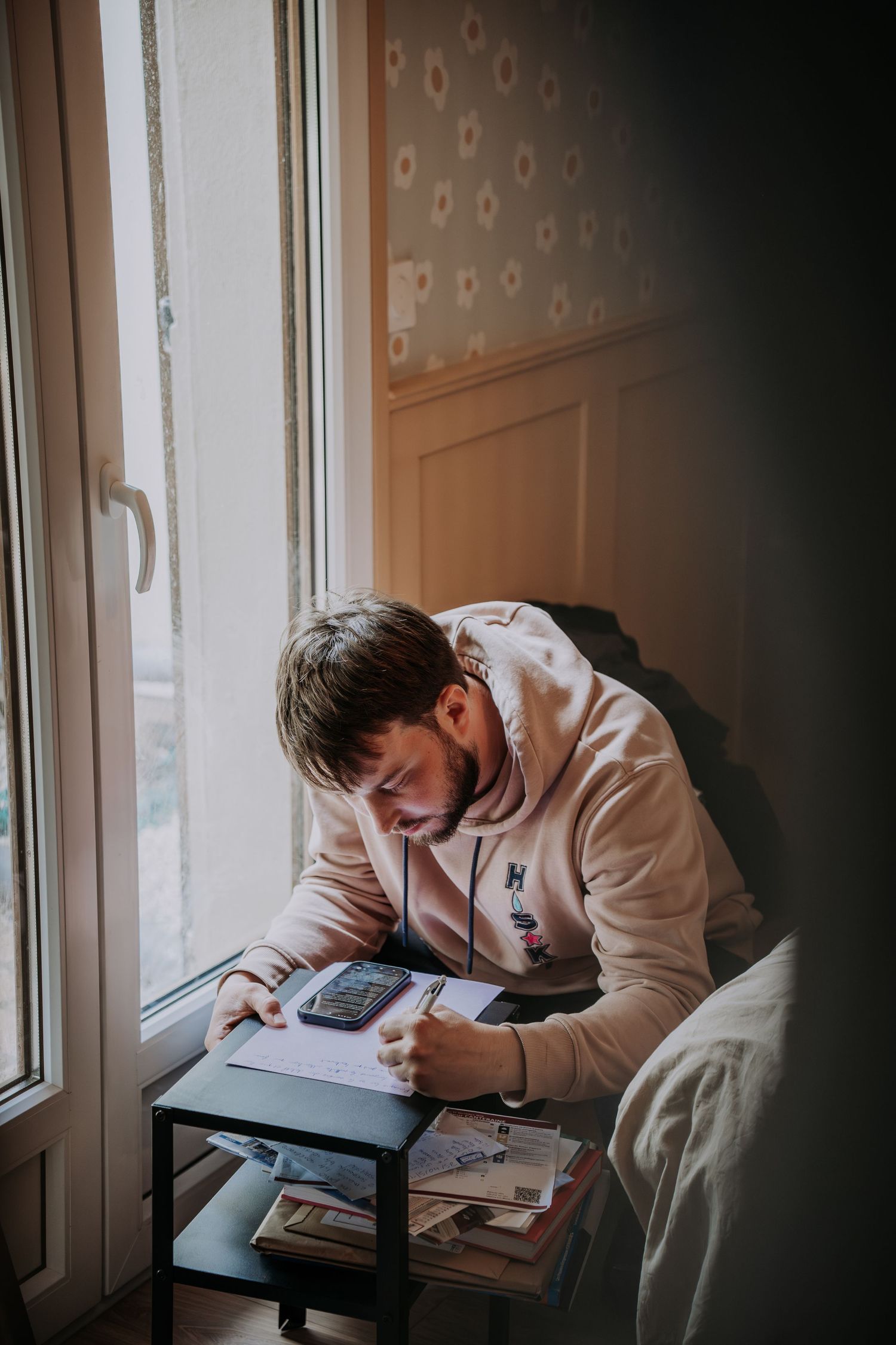 Young man in pink hoodie writing notes at small table by window with calculator nearby.