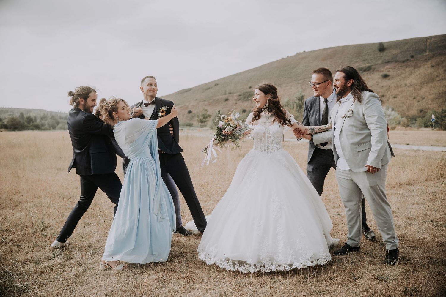 Wedding party celebrating outdoors in a field, bride in white gown with bridesmaids and groomsmen on a cloudy day.