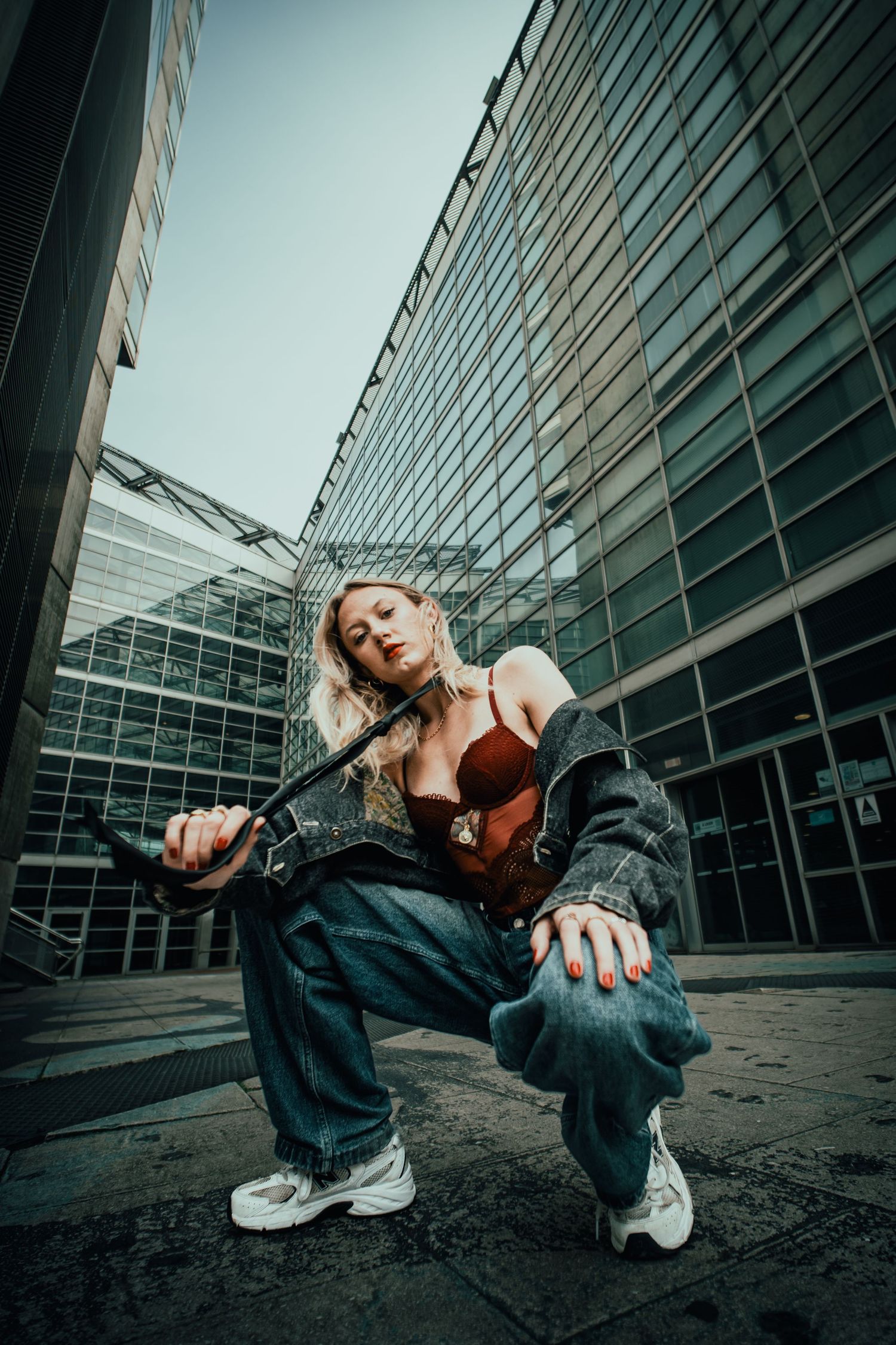 Woman in baggy jeans and leather jacket crouching confidently in front of modern glass building in urban setting.