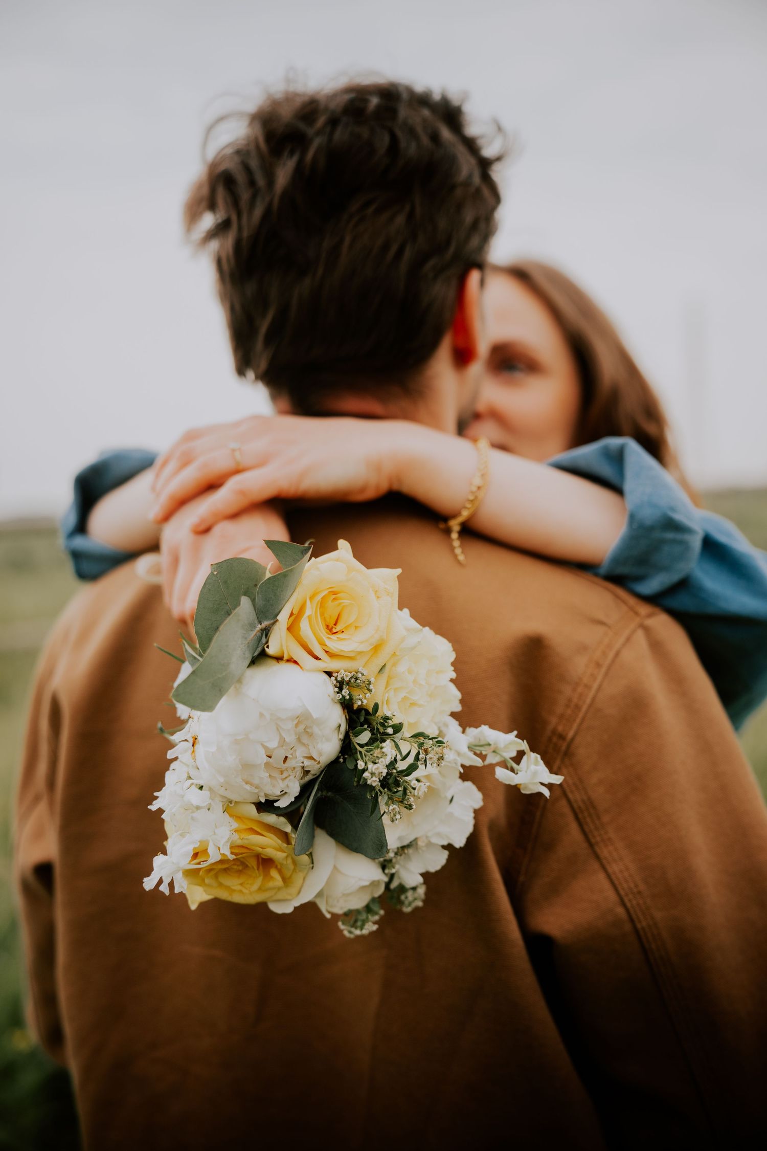 Couple embracing outdoors, woman holding a bouquet of yellow and white roses while hugging man in brown jacket.