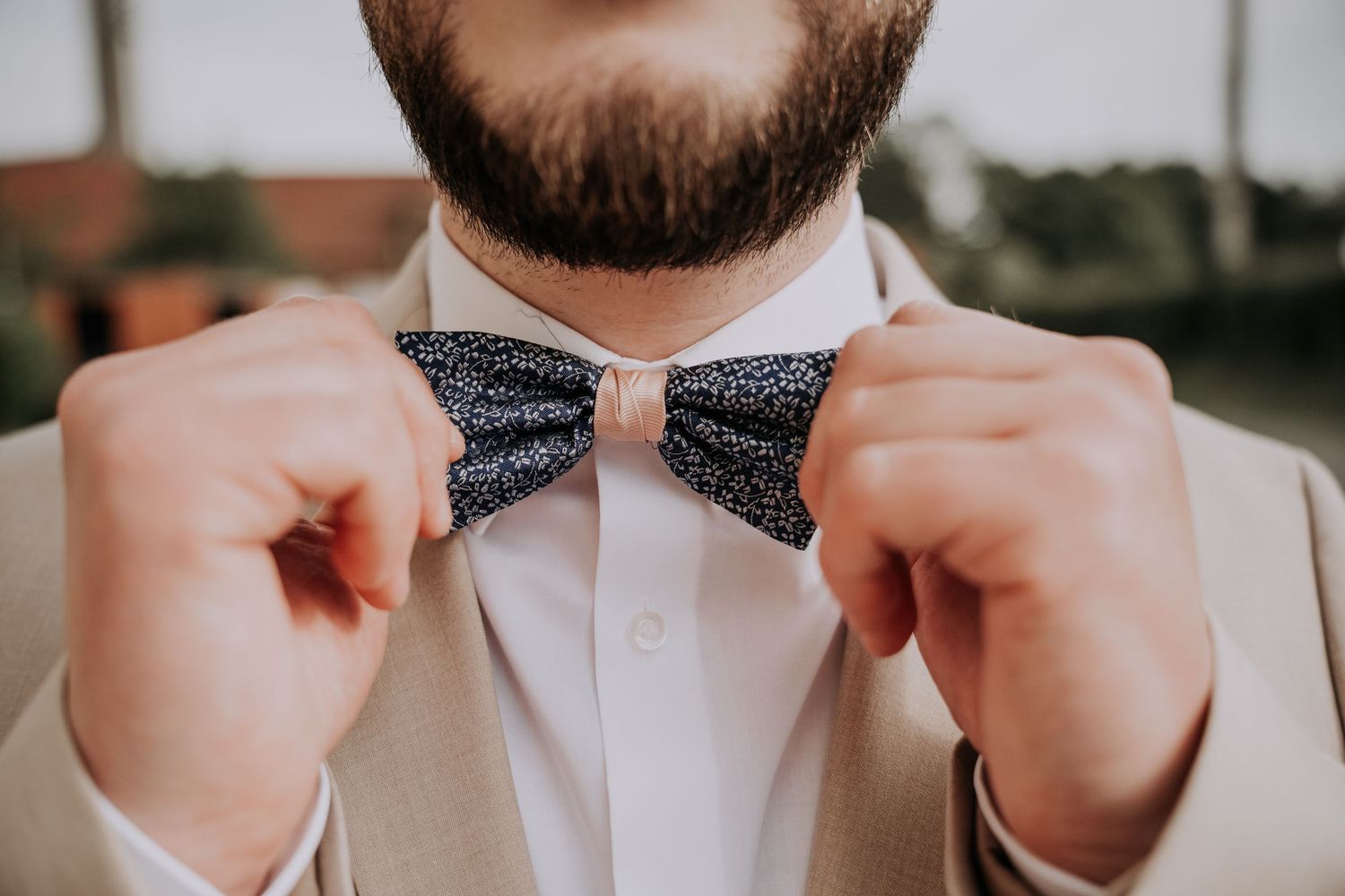 Bearded groom adjusting his navy blue patterned bow tie while wearing a beige suit and white dress shirt.