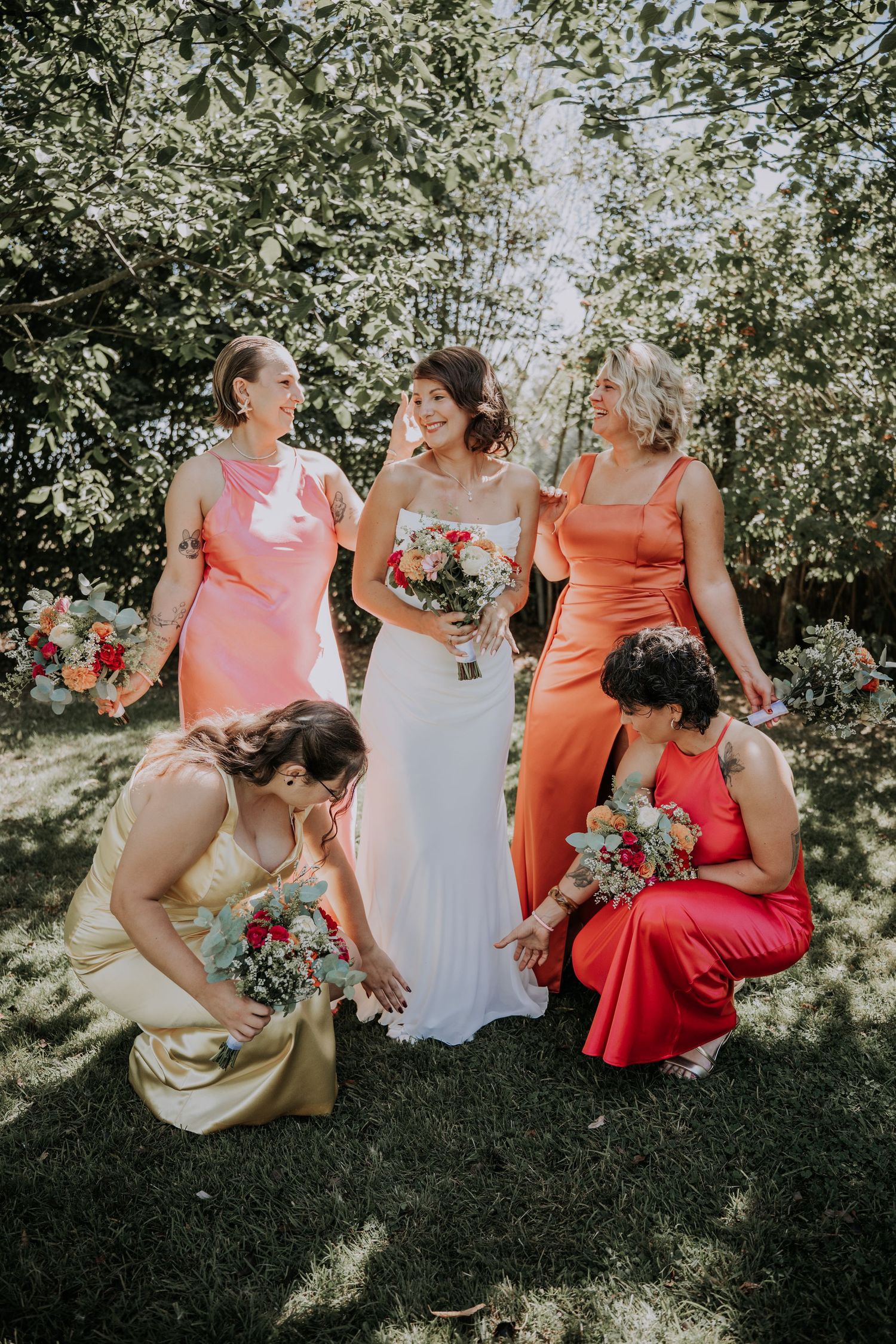 Bride in white gown poses with four bridesmaids in coral, orange, gold, and red dresses holding bouquets outdoors.