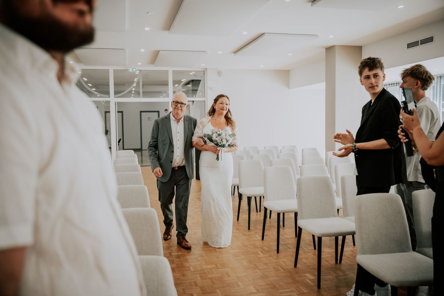 Bride walking down the aisle with her father in a modern wedding ceremony venue with white chairs.