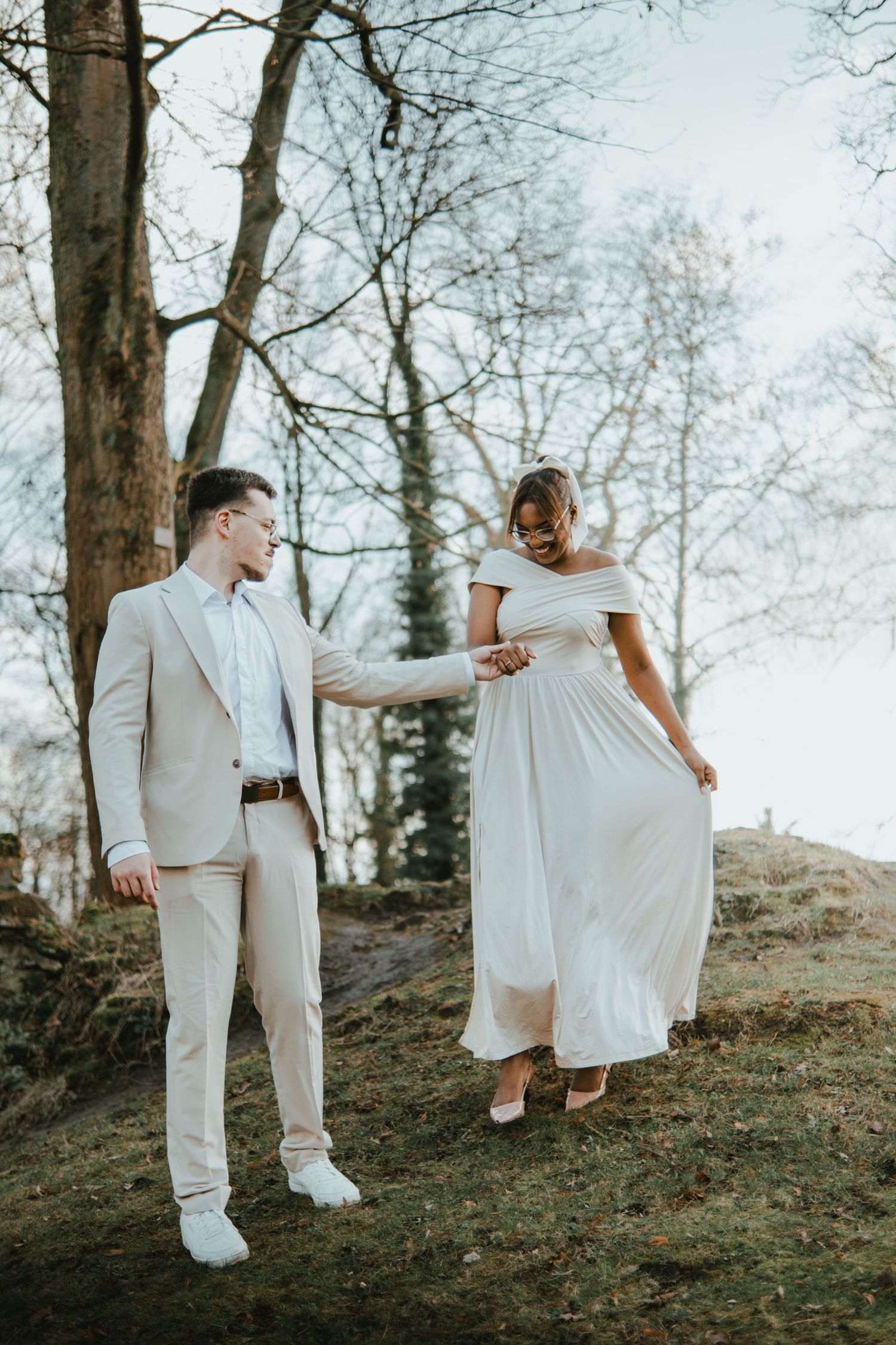 Couple in wedding attire holding hands outdoors, groom in beige suit, bride in white off-shoulder gown, among bare trees.