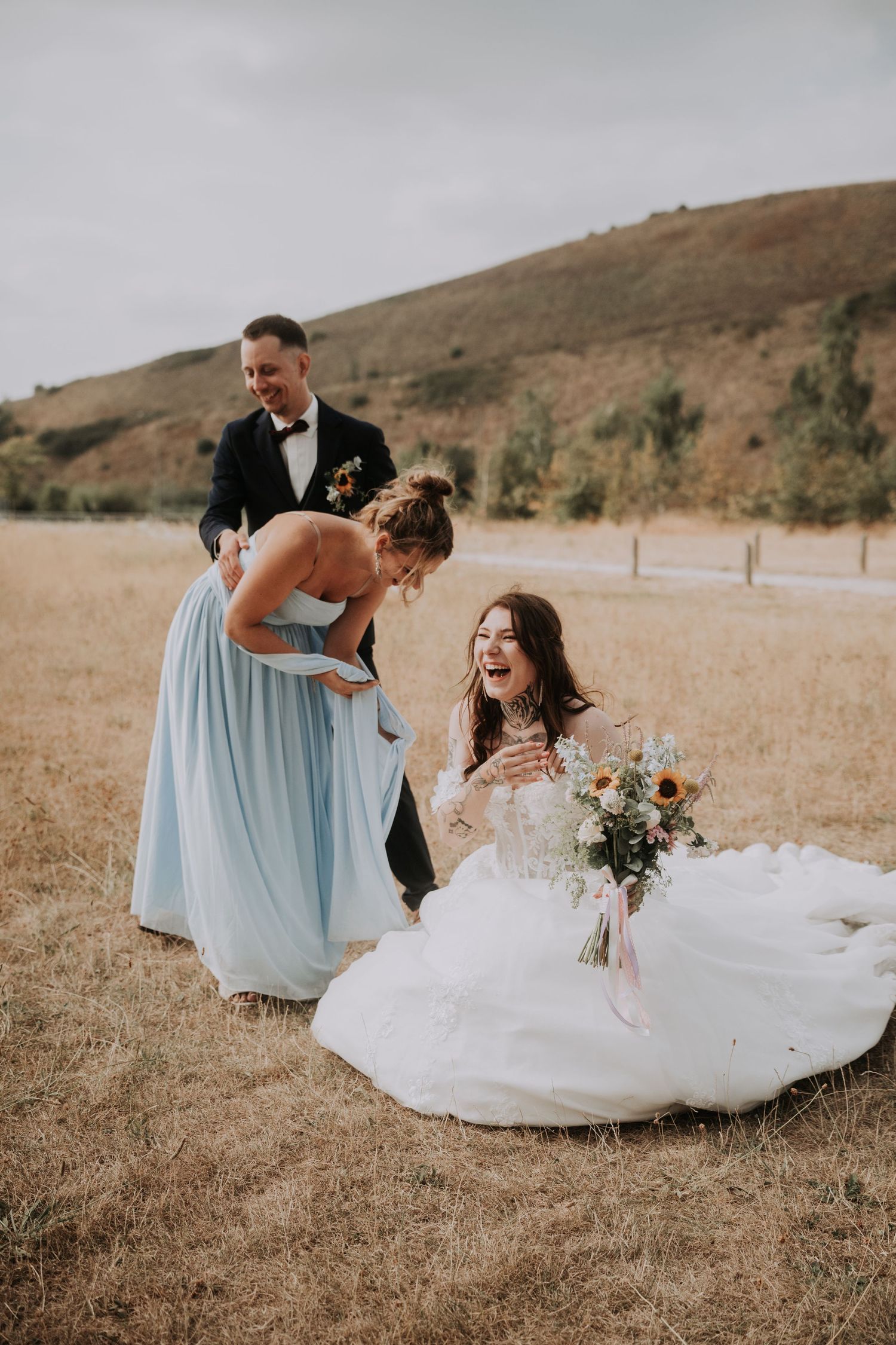 Bride laughing on ground in white gown with bridesmaid in blue dress and groom in tuxedo at outdoor wedding.