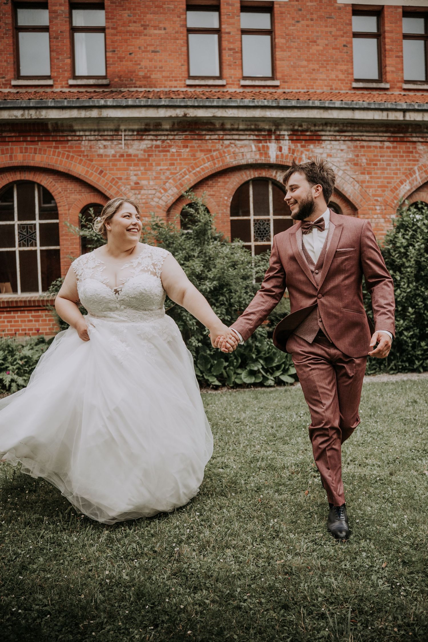 Bride in white gown and groom in burgundy suit hold hands, laughing while walking on grass before brick building.