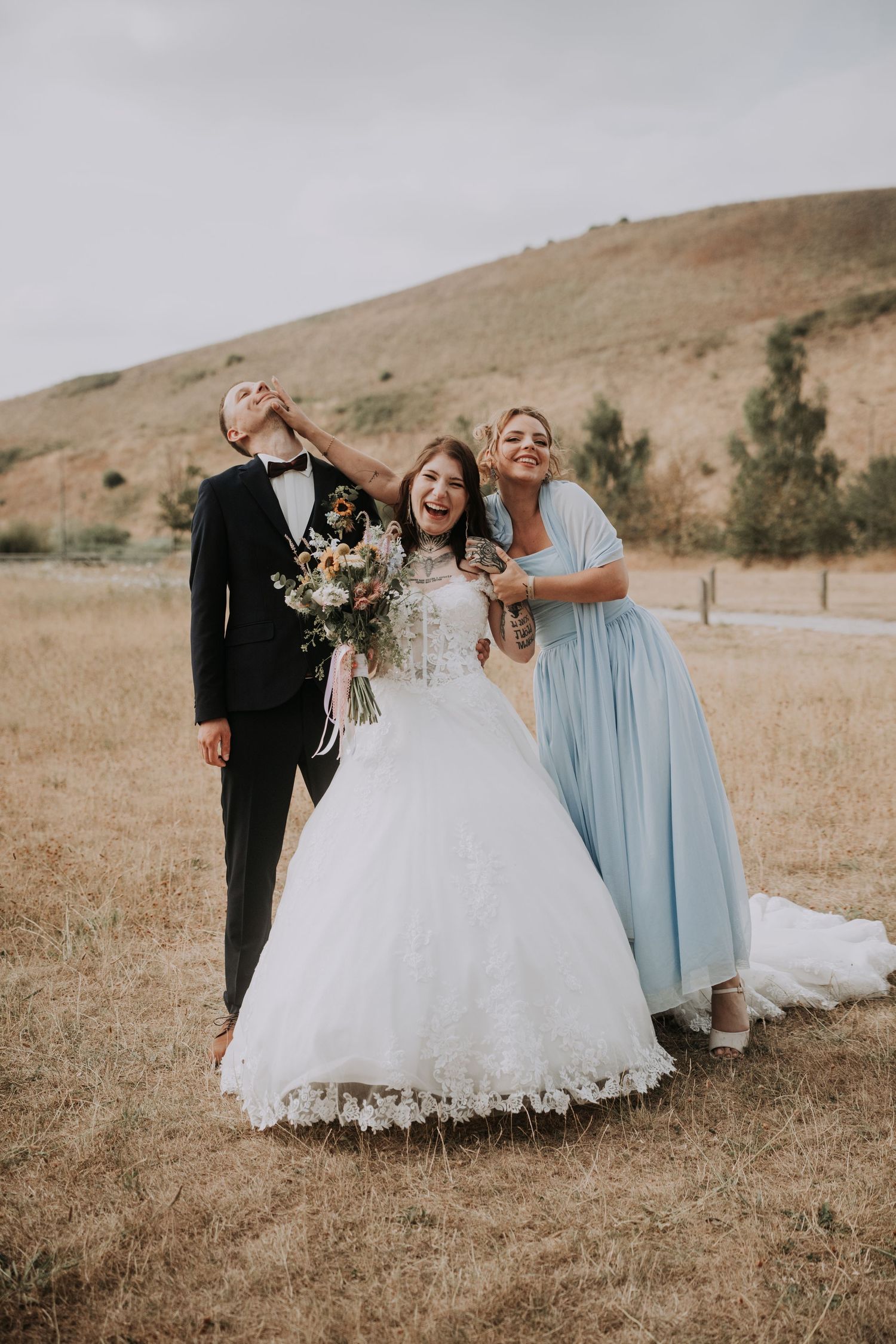 Bride in white gown and groom in tuxedo celebrate with bridesmaid in blue dress outdoors at rustic wedding.