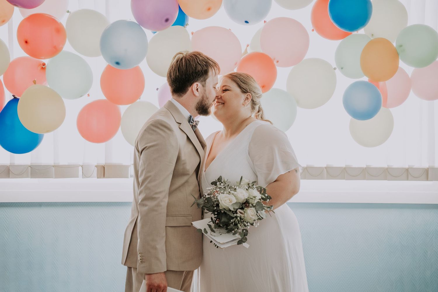 Bride and groom sharing a kiss in front of colorful balloon backdrop on their wedding day.