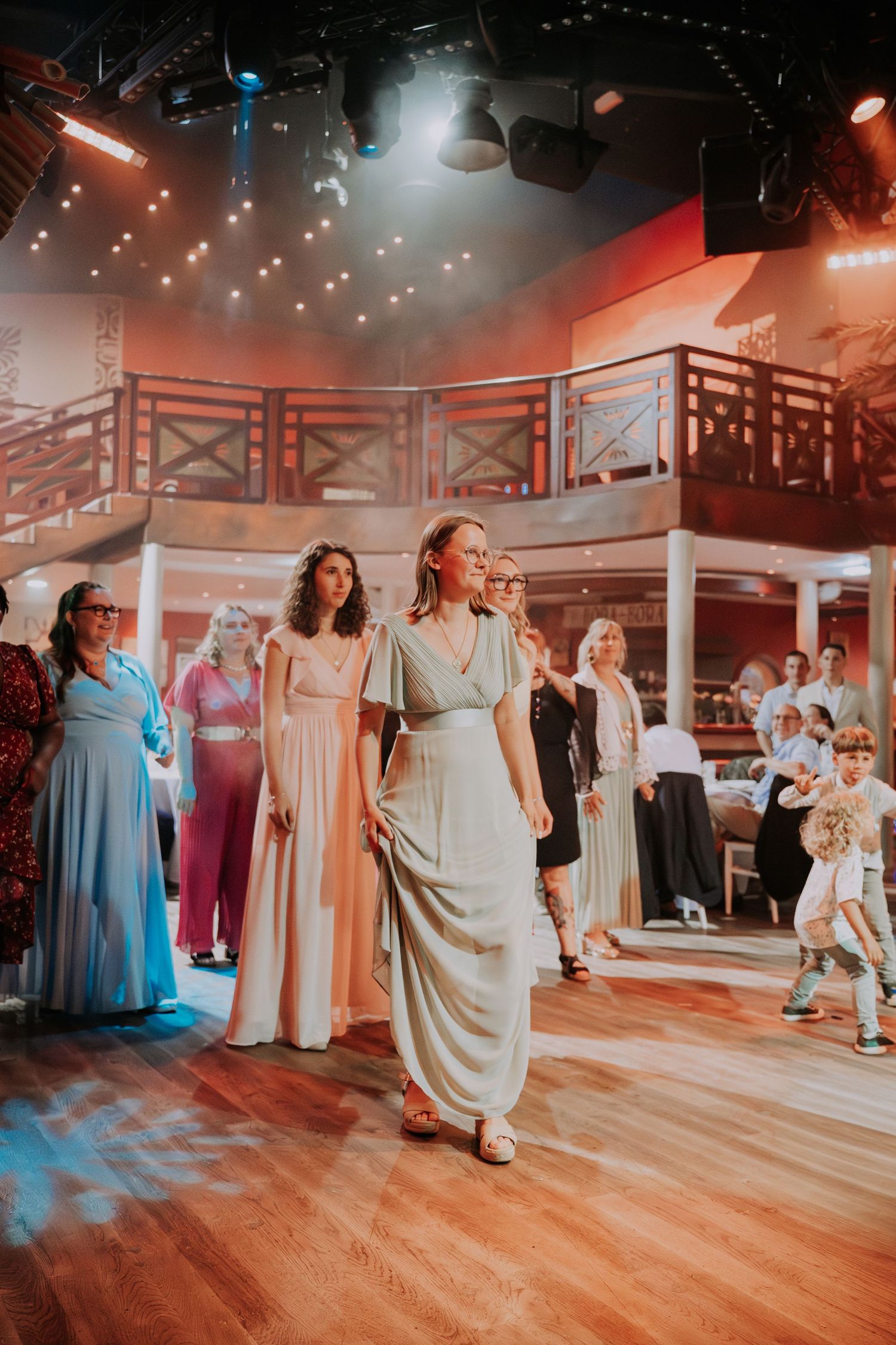 Women in elegant gowns walk a wooden stage floor in a warmly lit indoor venue with balcony backdrop.