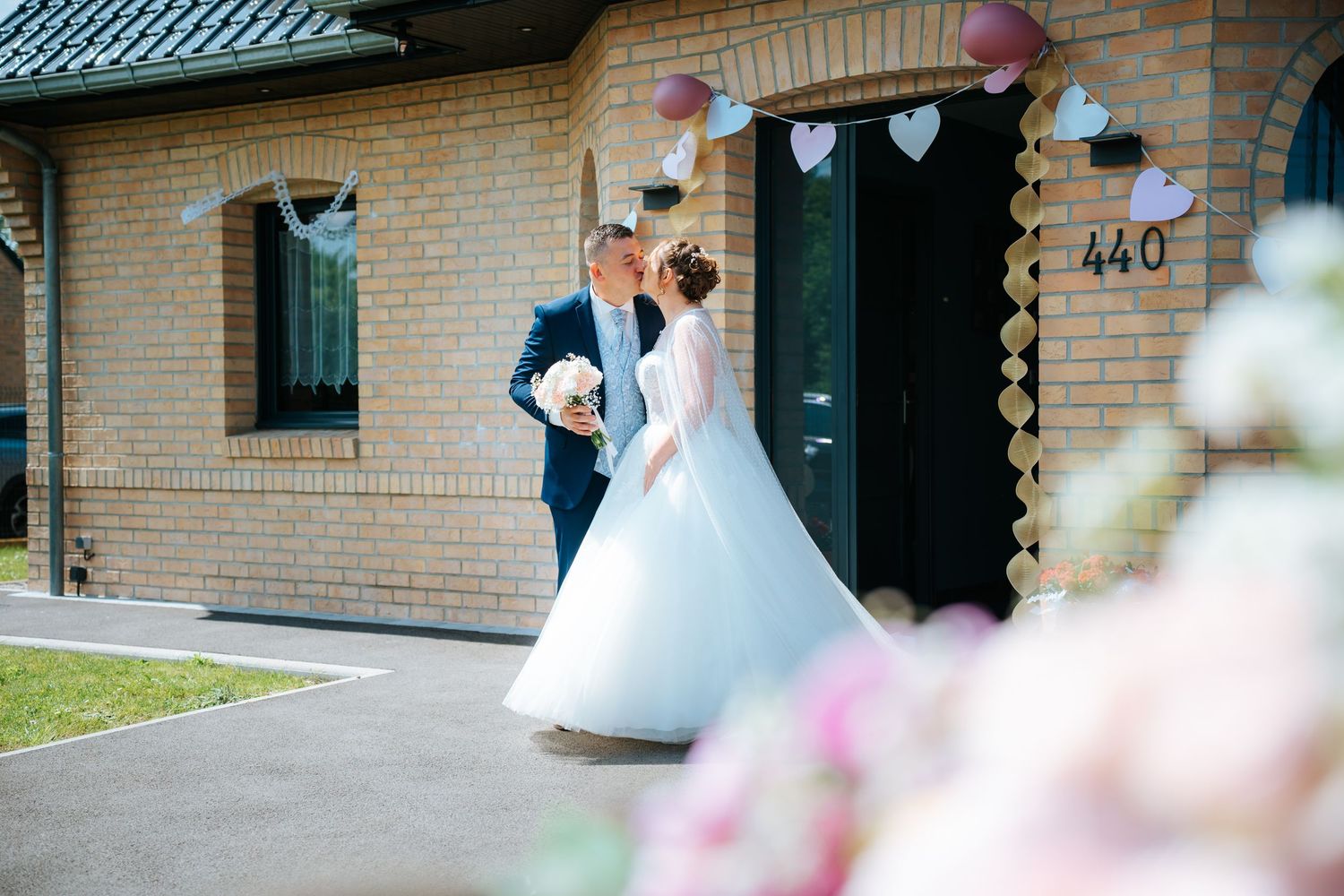 Bride and groom sharing a kiss outside a brick building decorated with heart bunting at their wedding.