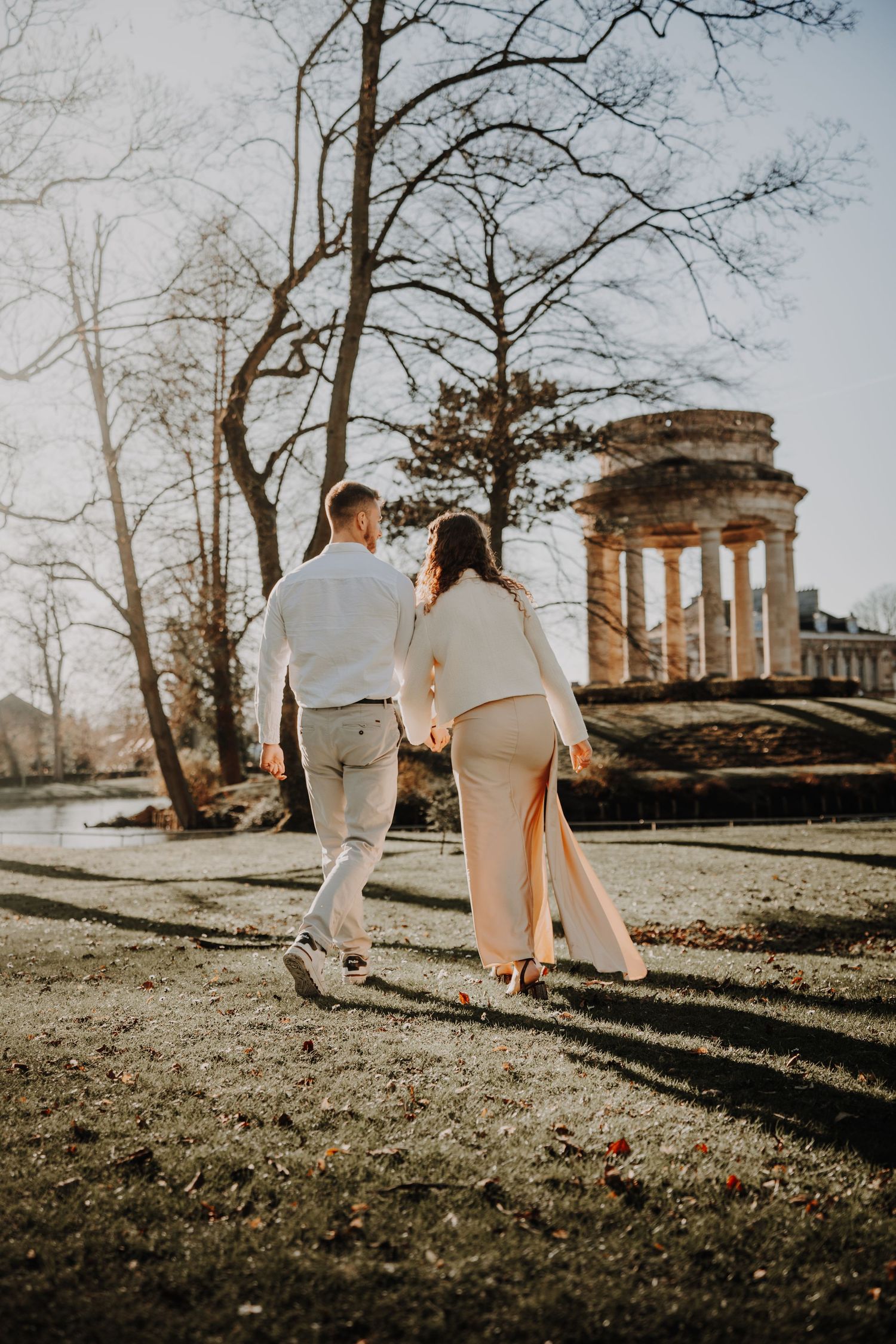 Couple holding hands walking toward a classical stone temple in a sunlit park during winter.