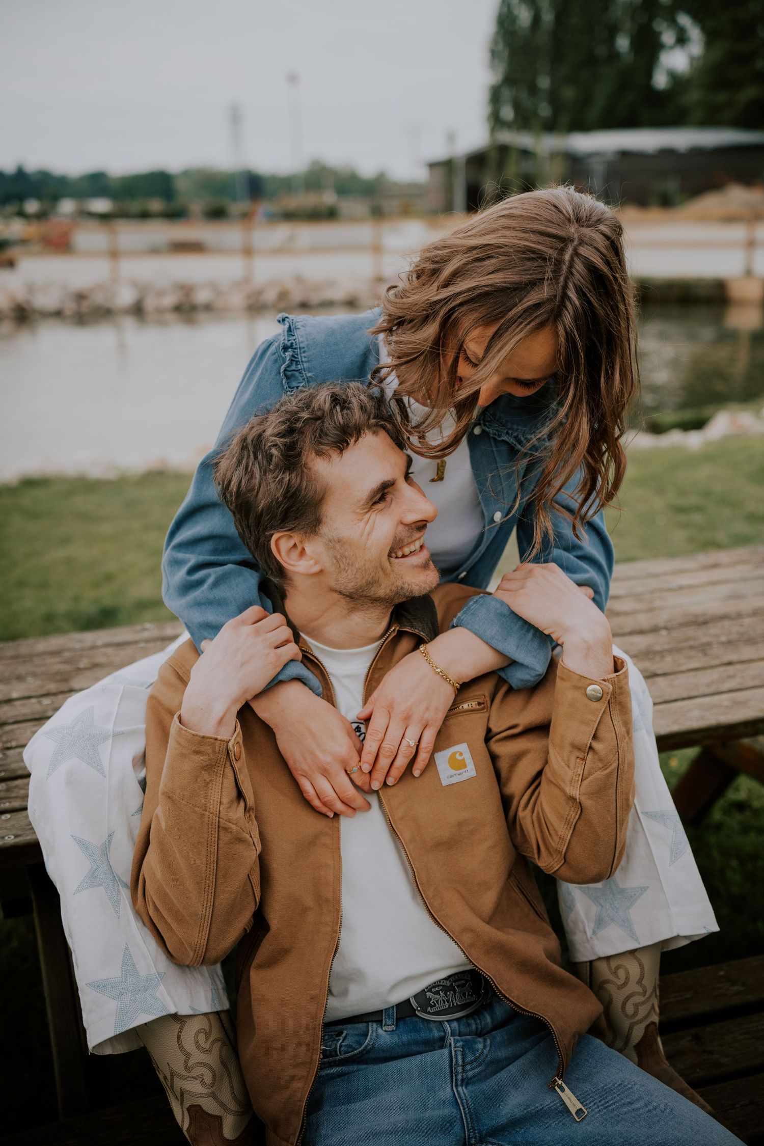 A smiling couple sharing a tender moment outdoors, woman in denim jacket leaning over man in Carhartt coat by a lake.