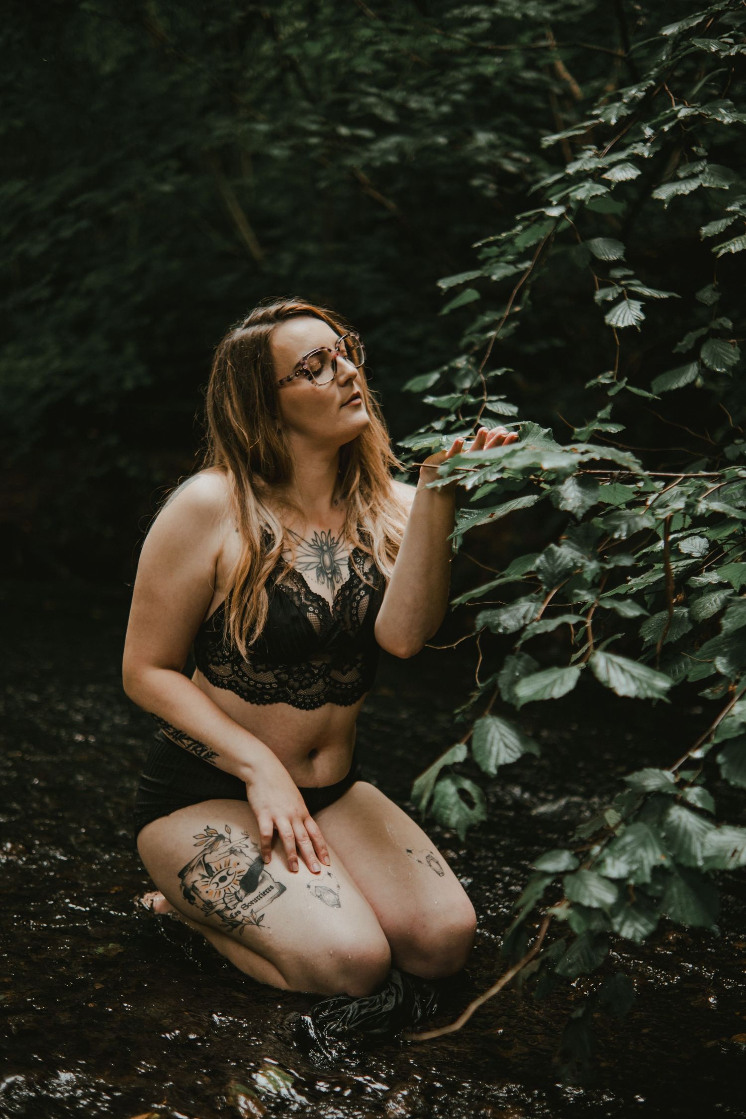Tattooed woman in black lace lingerie kneeling in a forest stream surrounded by lush green foliage.