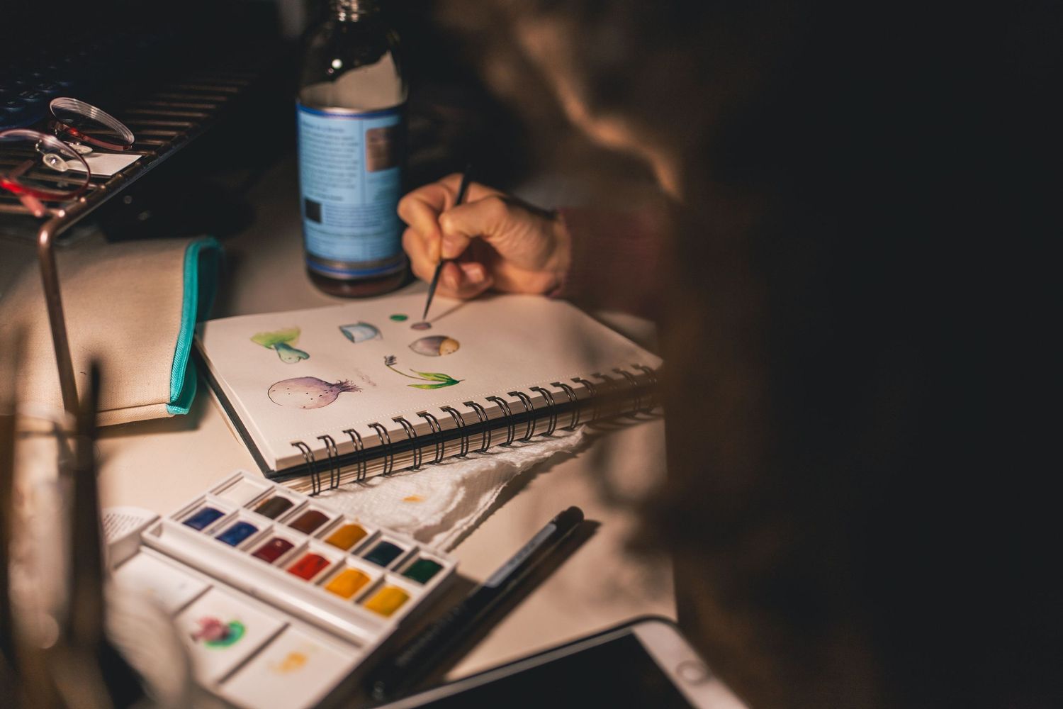 Artist painting colorful watercolor sketches in a spiral notebook with a paint palette on a dimly lit table.