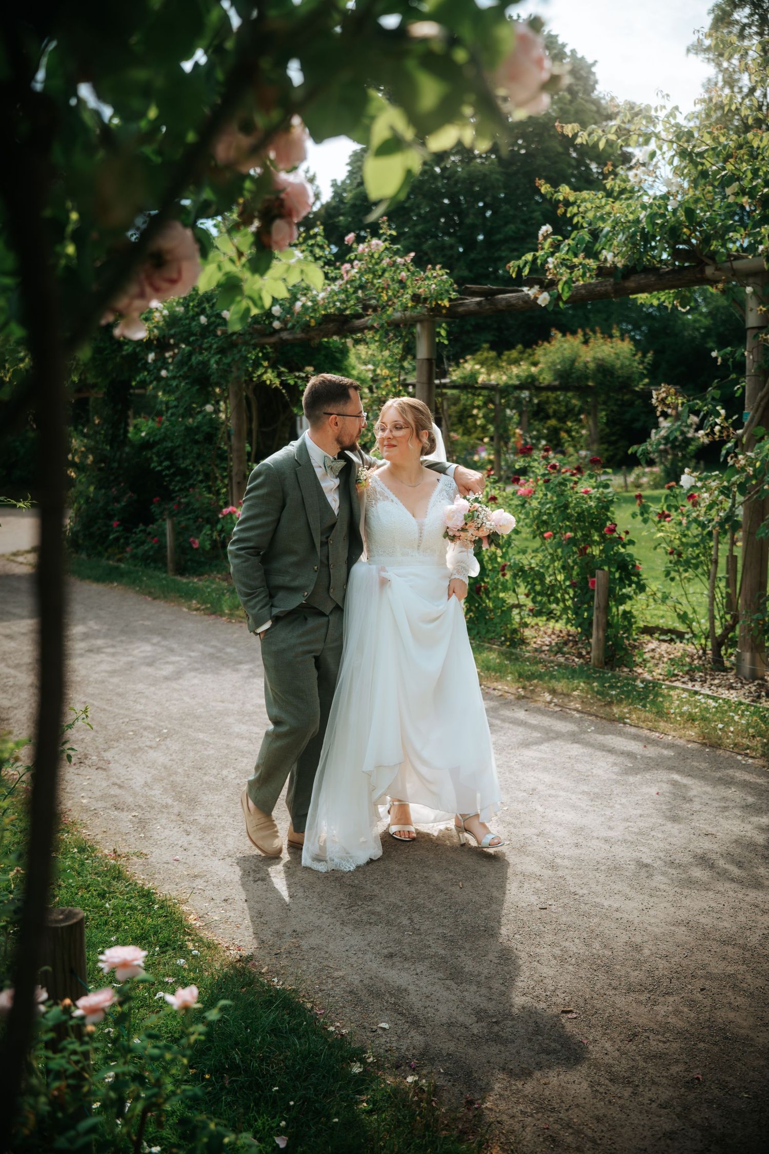 Bride and groom walking hand in hand through a lush garden path surrounded by blooming roses on their wedding day.