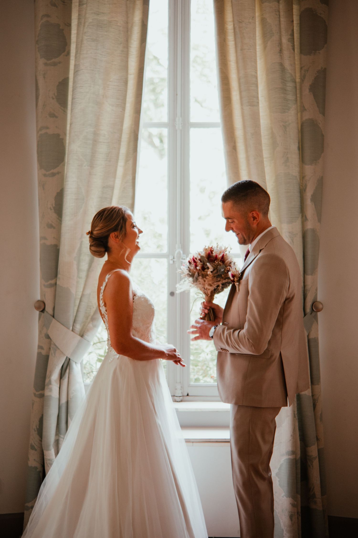 Bride and groom share a tender moment by a sunlit window, with the groom holding a bouquet.