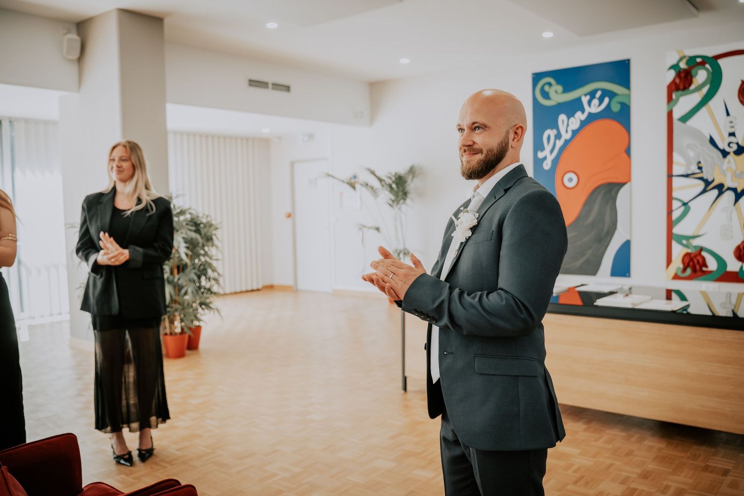 Bald bearded man in dark suit clapping hands smiling indoors, woman in black dress and colorful artwork in background.