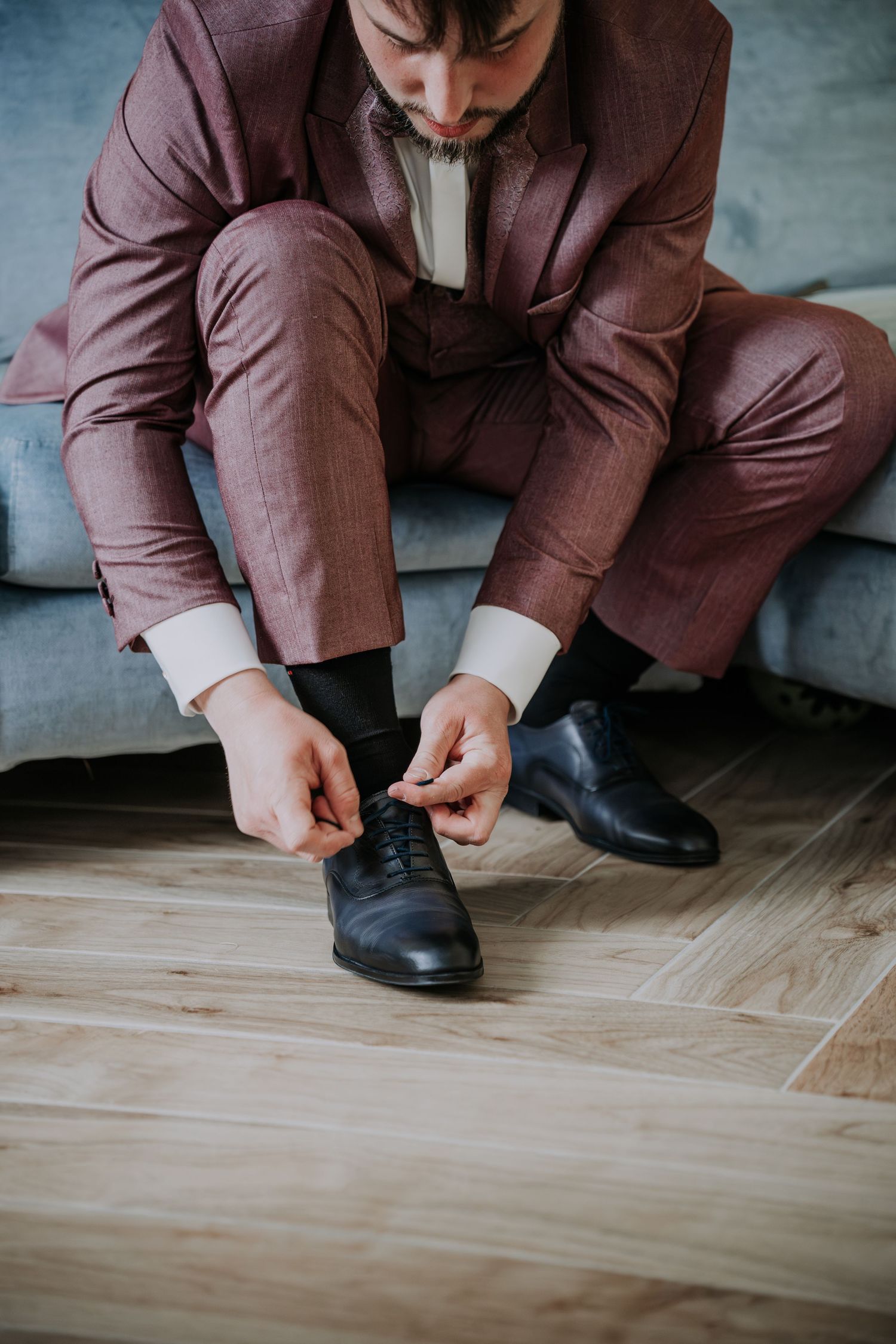 Man in burgundy suit tying black dress shoes while seated on a gray sofa on hardwood floor.