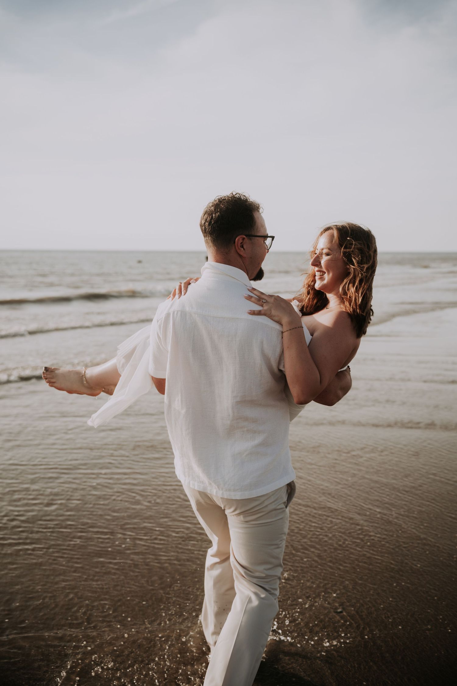 A groom carries his smiling bride on a beach, both dressed in white, with ocean waves in the background.