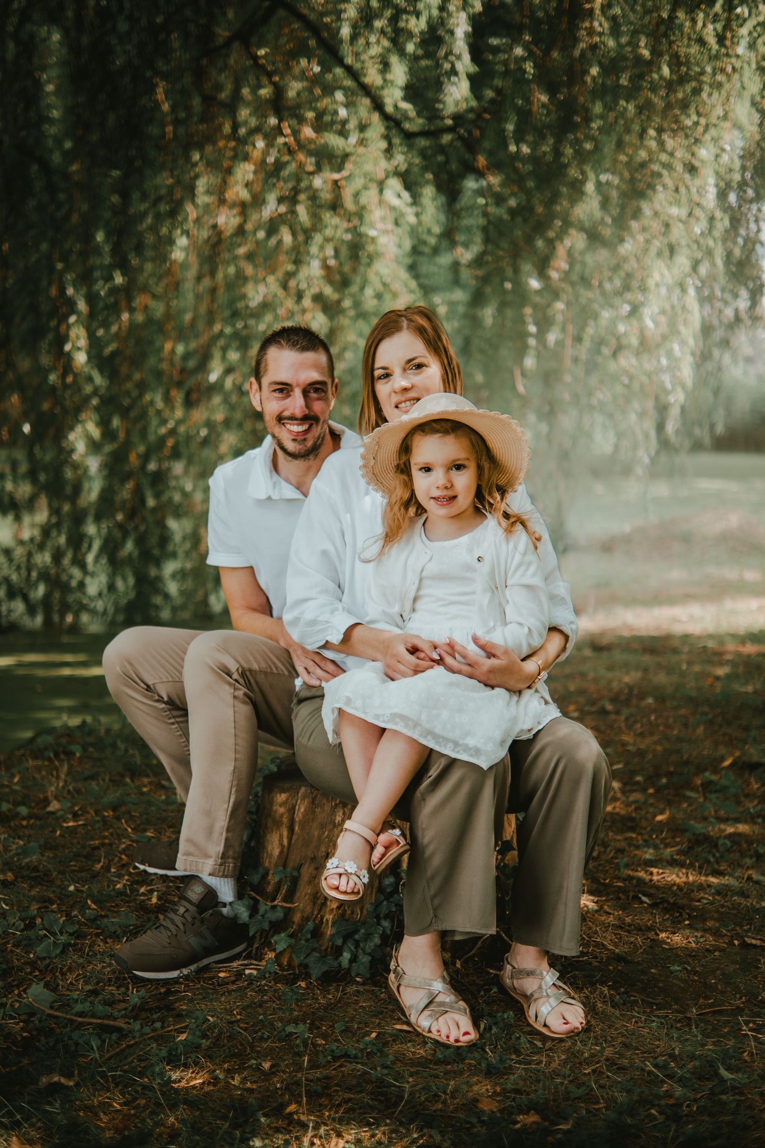 Happy family of three posing outdoors under a weeping willow tree, parents and young daughter in white outfits.