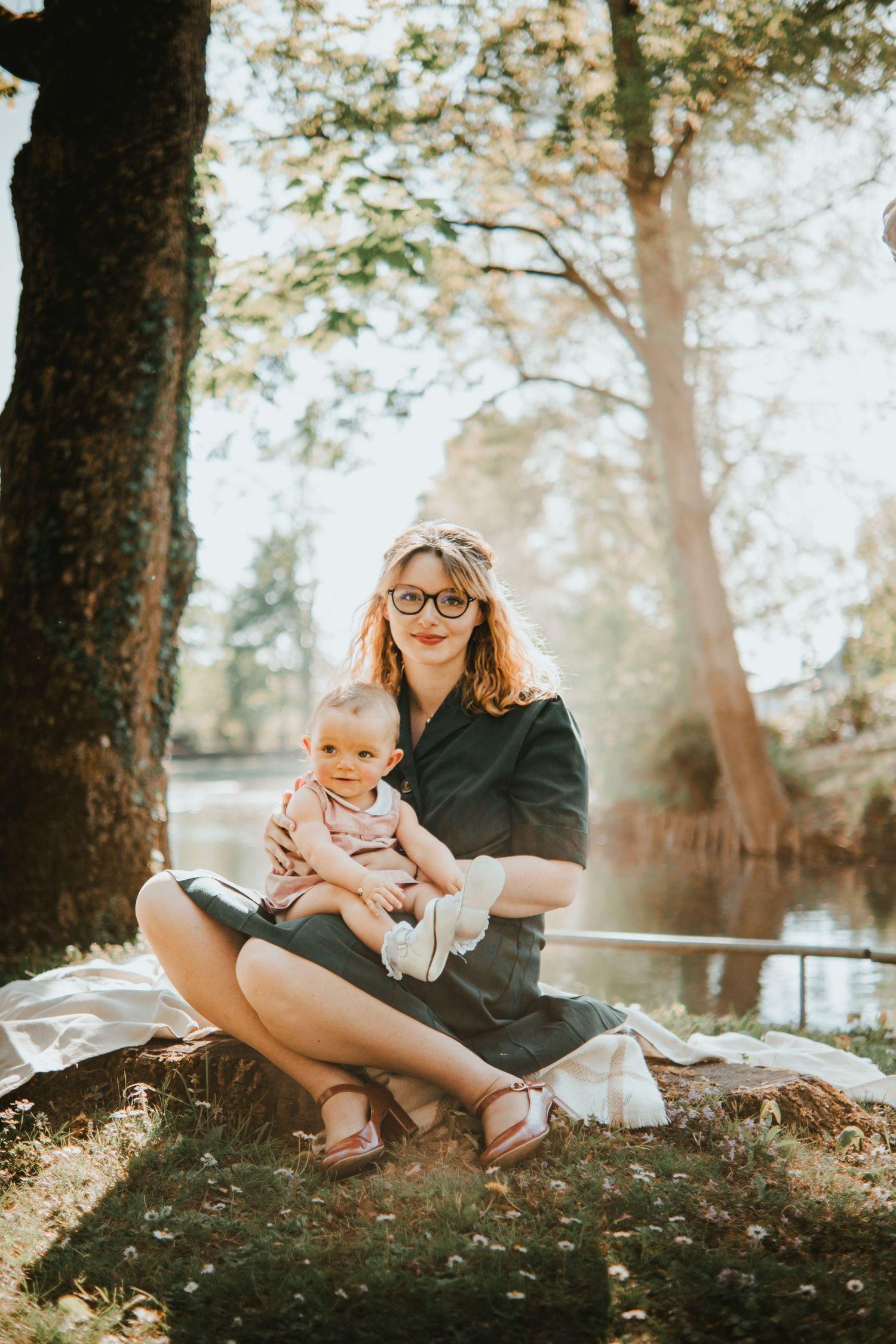 A young mother with glasses sits cross-legged on grass holding her baby near a sunlit pond surrounded by trees.