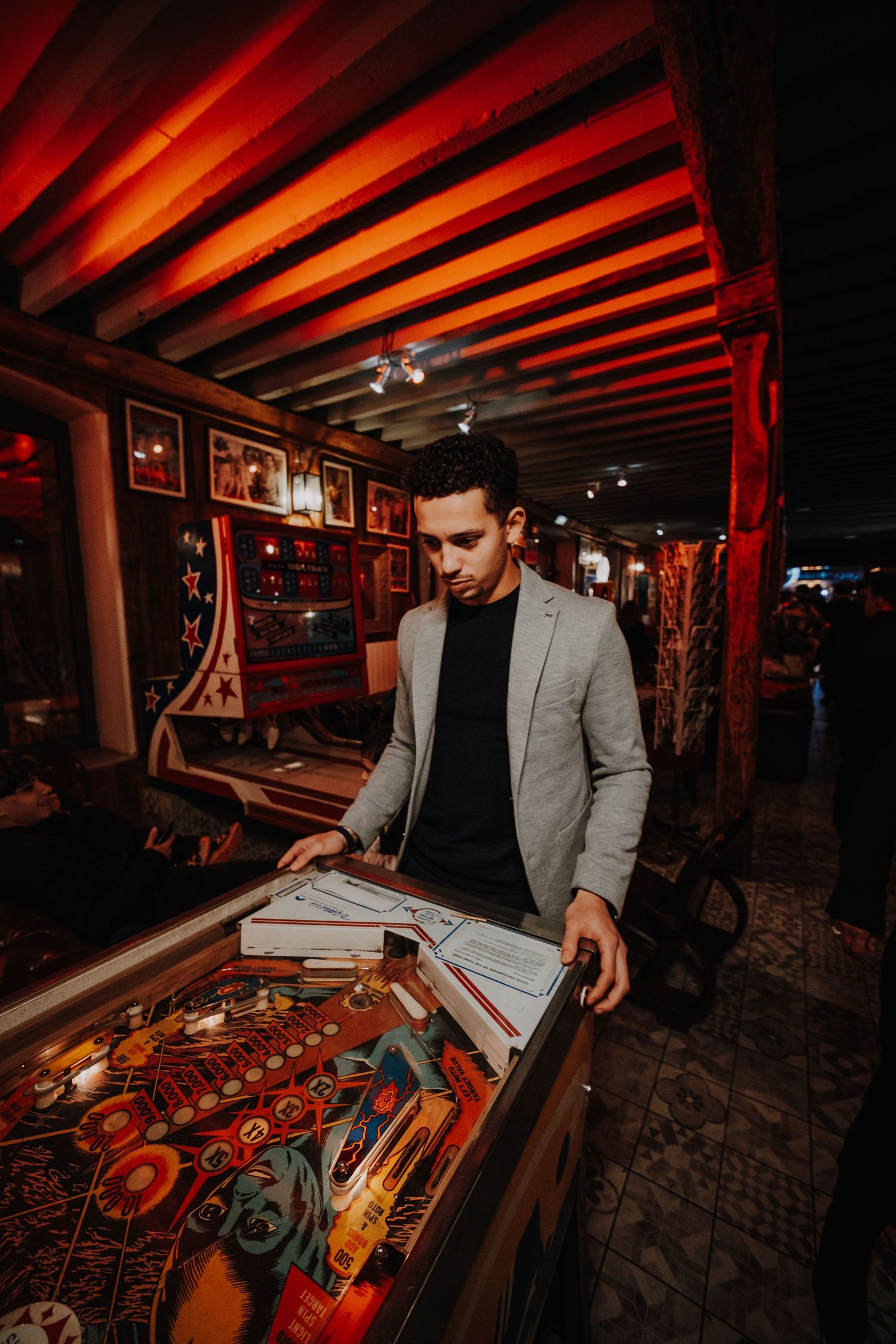 Man in gray blazer playing pinball machine in dimly lit arcade bar with red lighting.