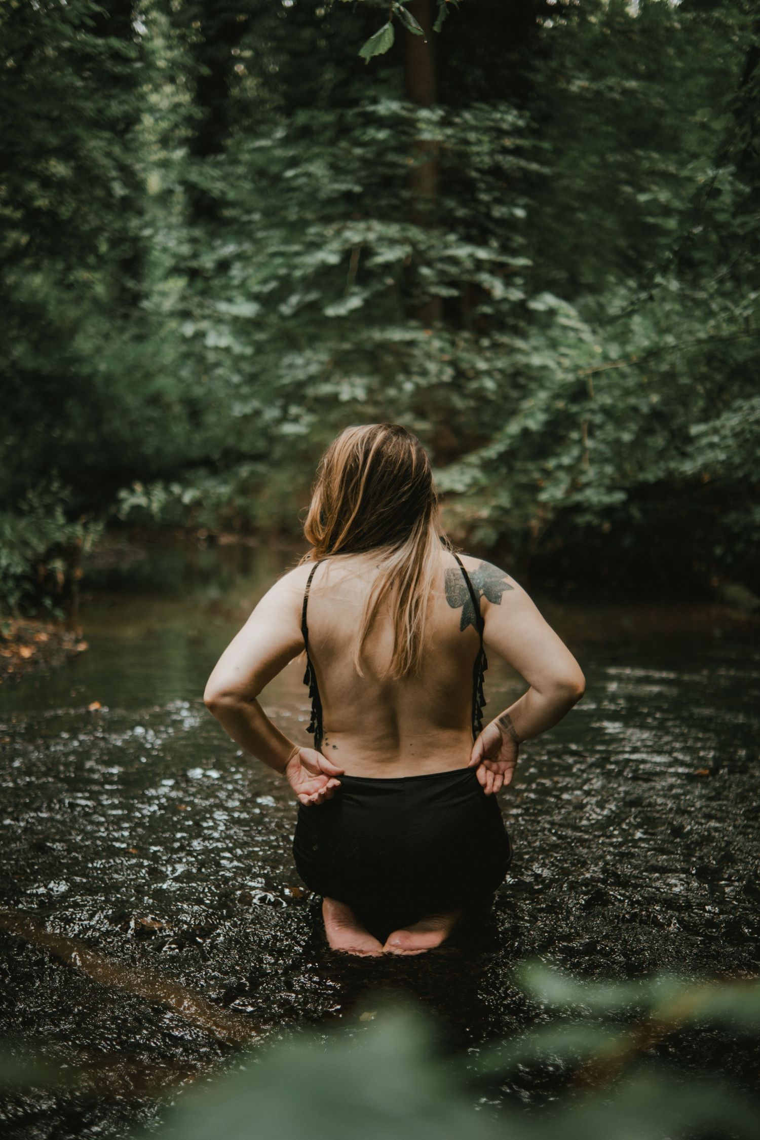 Woman with tattoo standing in a forest creek wearing black swimwear, surrounded by lush green trees.