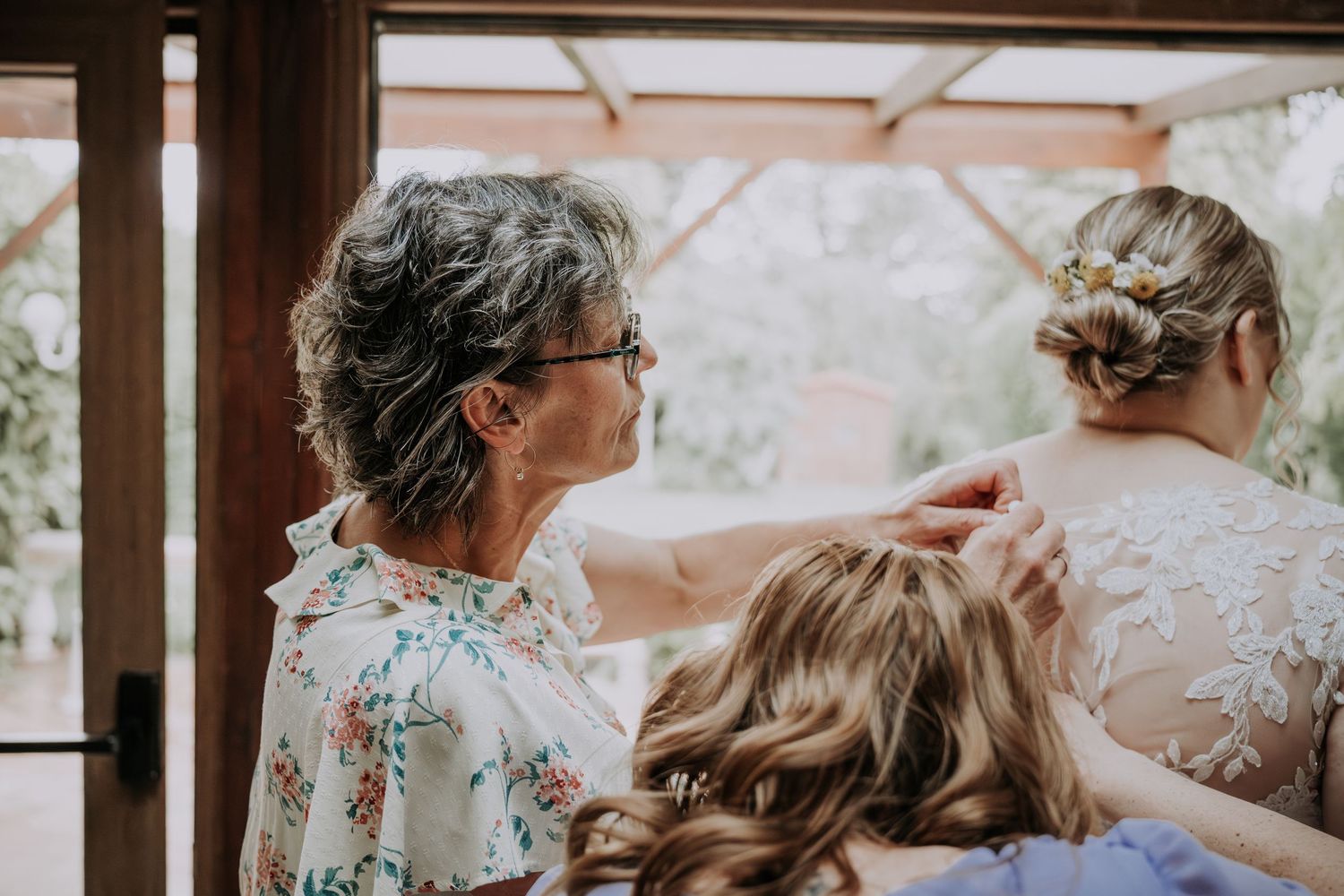 Older woman in floral dress helping style bride's hair during wedding preparations indoors.