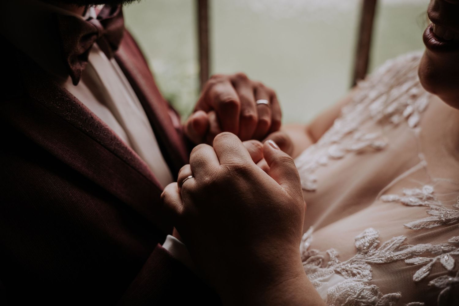 Close-up of bride and groom holding hands, showcasing wedding rings, with lace bridal gown and burgundy suit details.