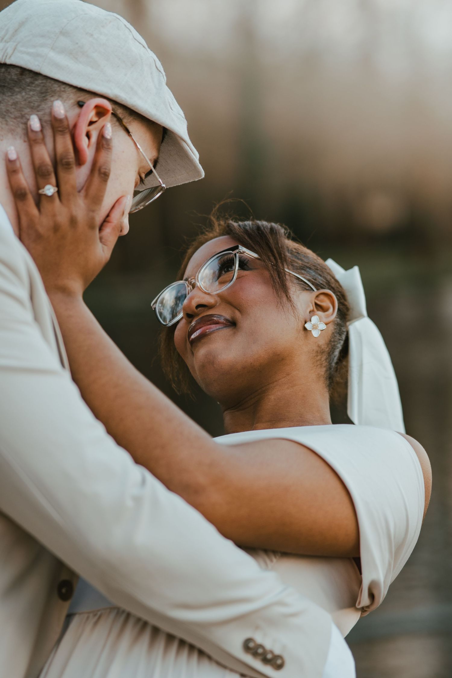 A couple in cream wedding attire shares a romantic embrace, the bride smiling up at her partner outdoors.