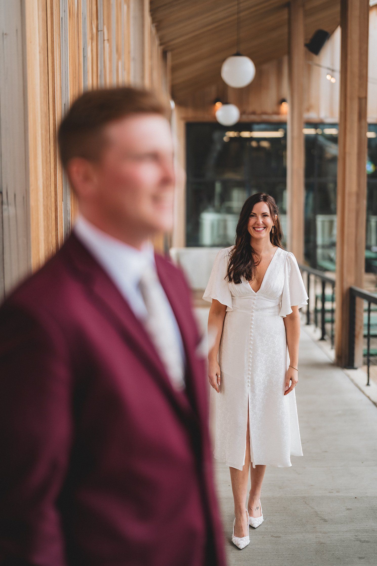 Smiling bride in white midi dress approaches groom in burgundy suit along a modern wooden corridor.