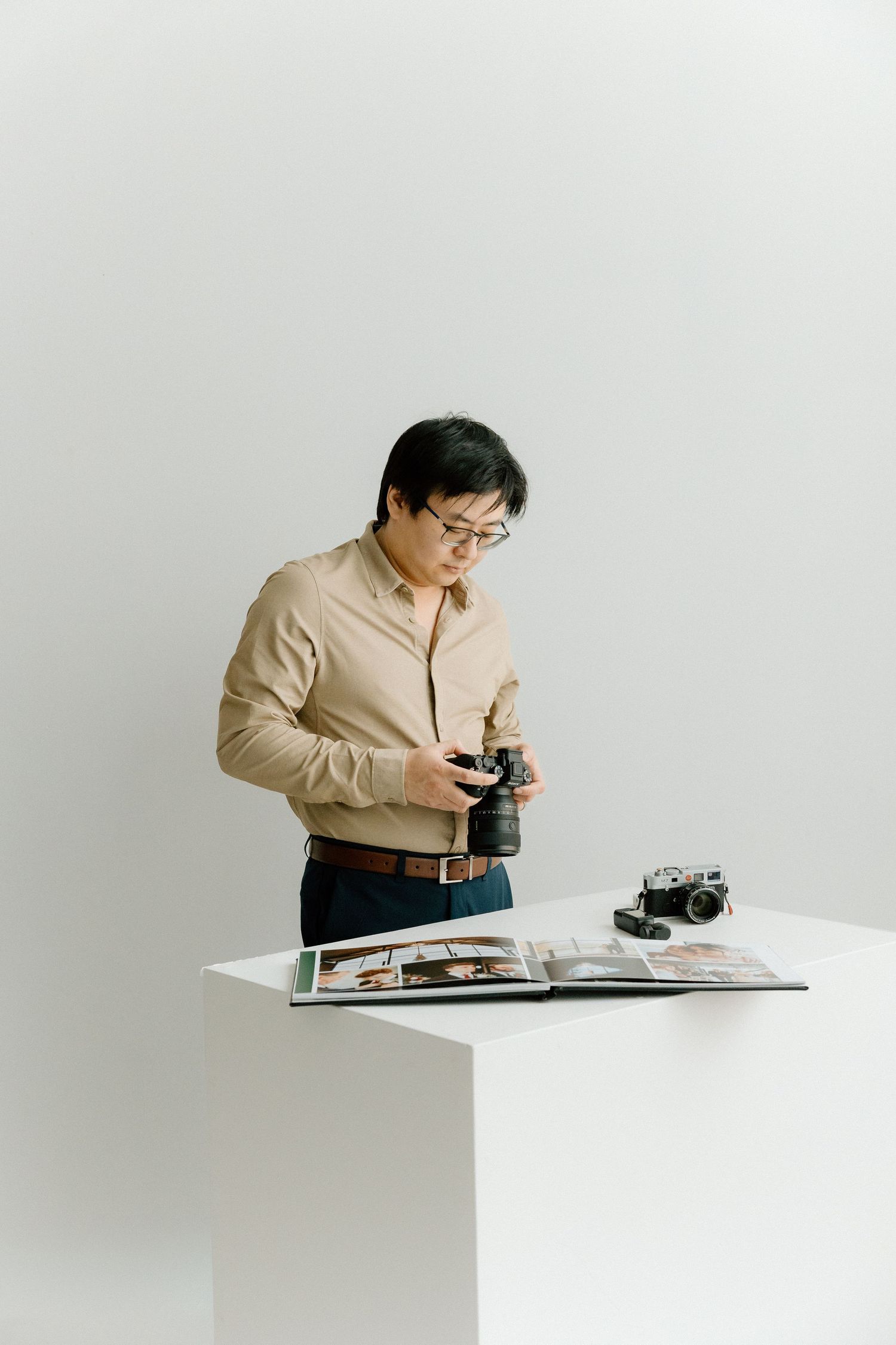A person in a beige shirt examines a vintage camera while standing at a white table in a minimalist setting.