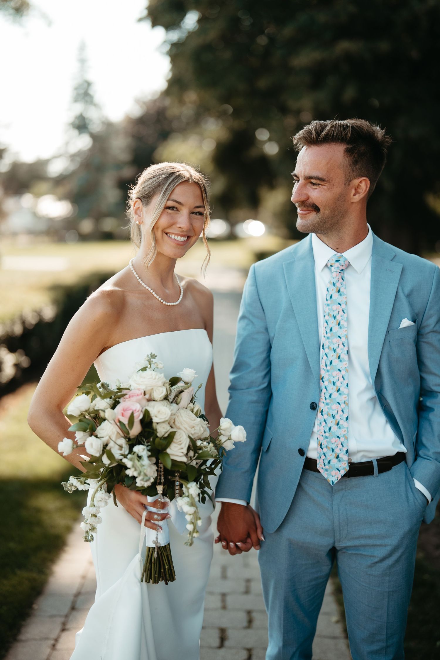 Smiling bride in white strapless gown holds floral bouquet beside groom in light blue suit on sunny garden path.