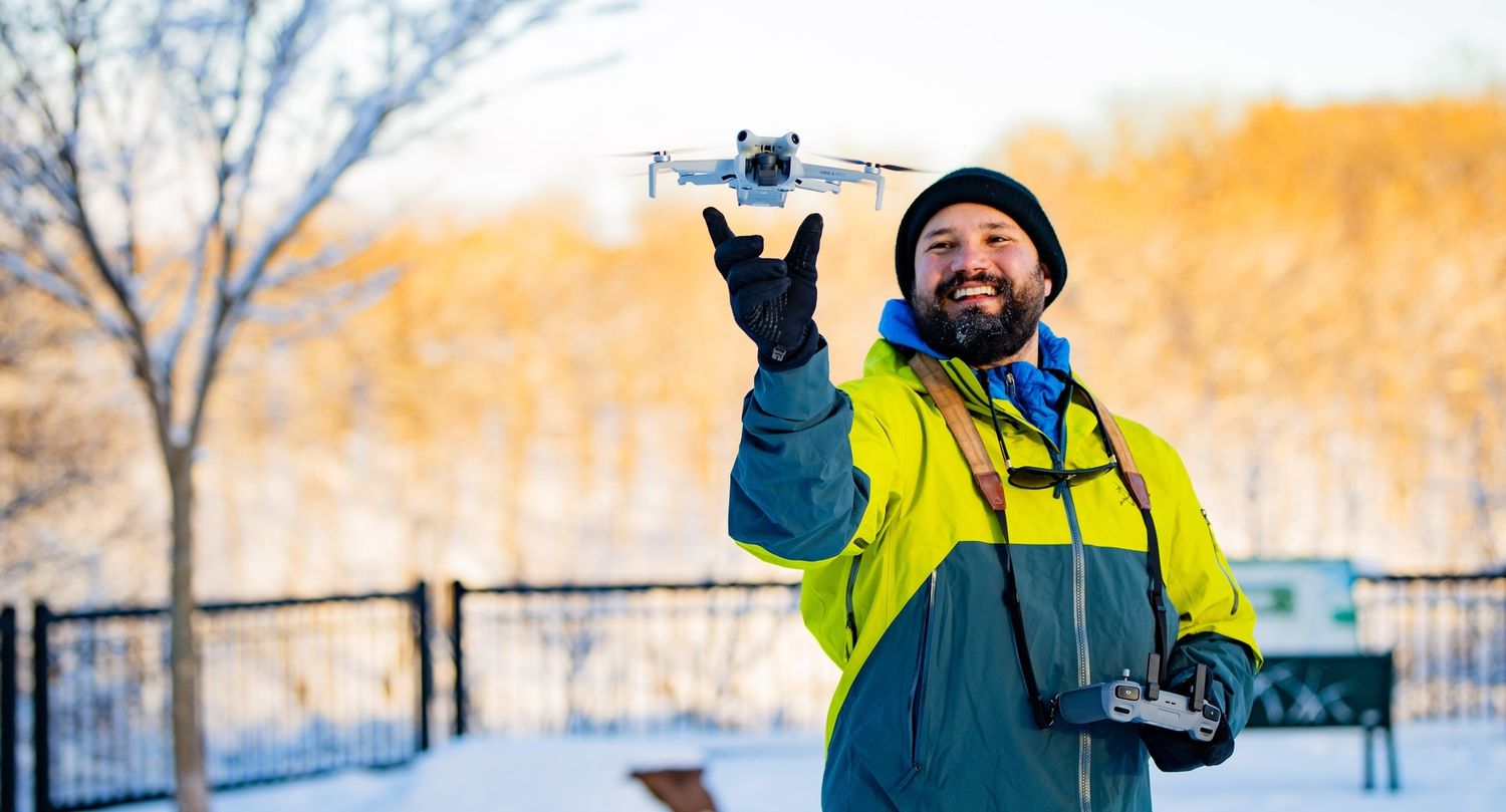 A person in winter gear holds up a drone while standing in a snowy landscape with trees in the background.